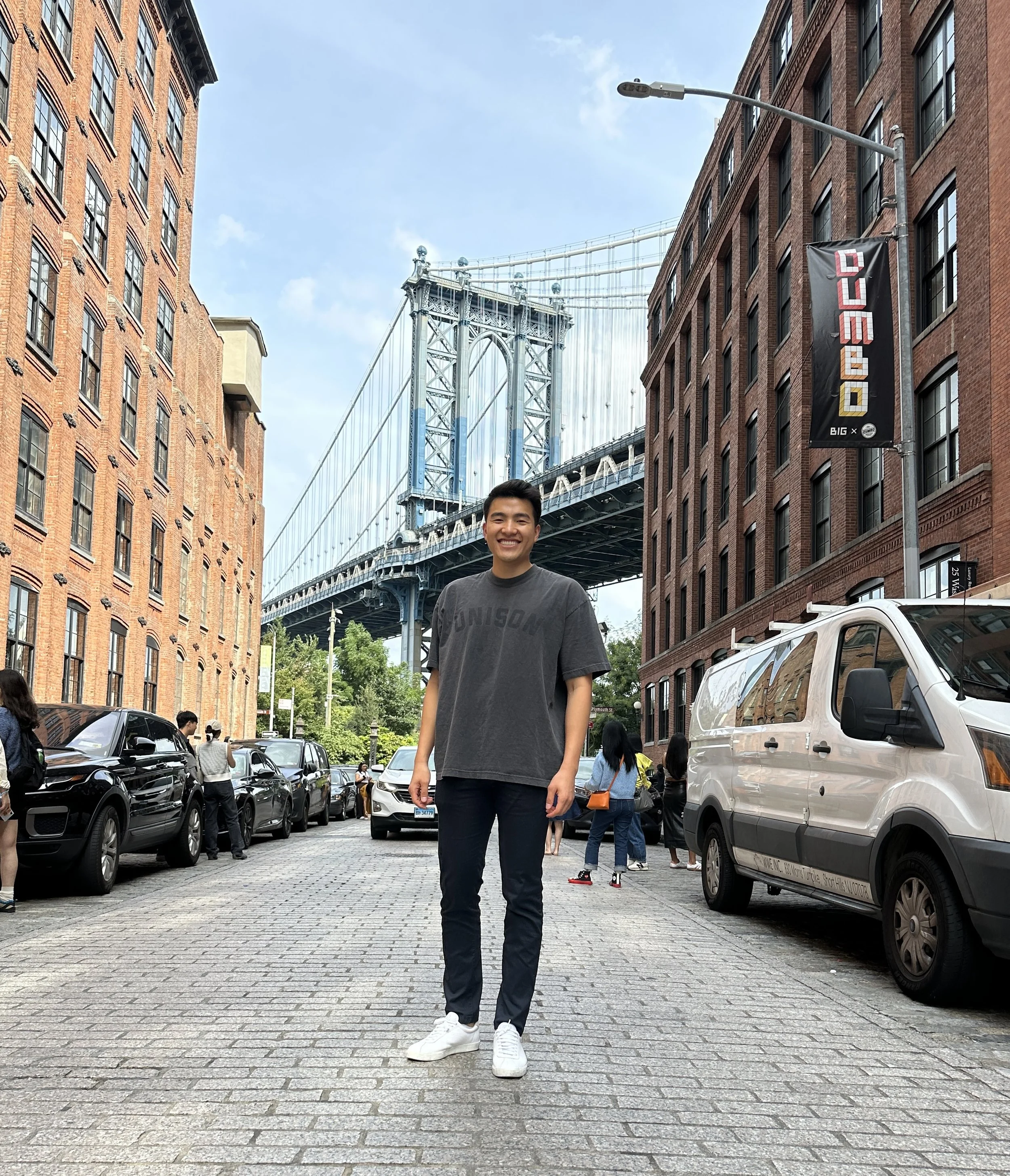 Young man smiling and standing on a cobblestone street in Dumbo, Brooklyn, with the Manhattan Bridge in the background. Brick buildings and parked cars line the street, and a banner reading 'DUMBO' is visible.