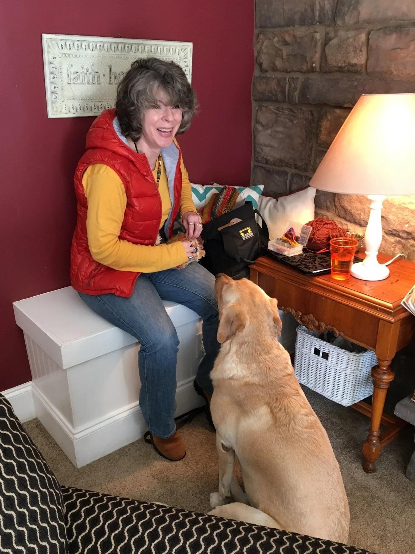 A woman sitting on a white storage bench smiling at a large Labrador Retriever dog that is sitting on the carpet and looking up at her. The woman wears a yellow shirt, a red vest, and jeans. There is a wooden side table with a lamp, a glass of what appears to be water, and various small items. The wall behind them is painted red with a decorative sign, and there is a stone wall section.