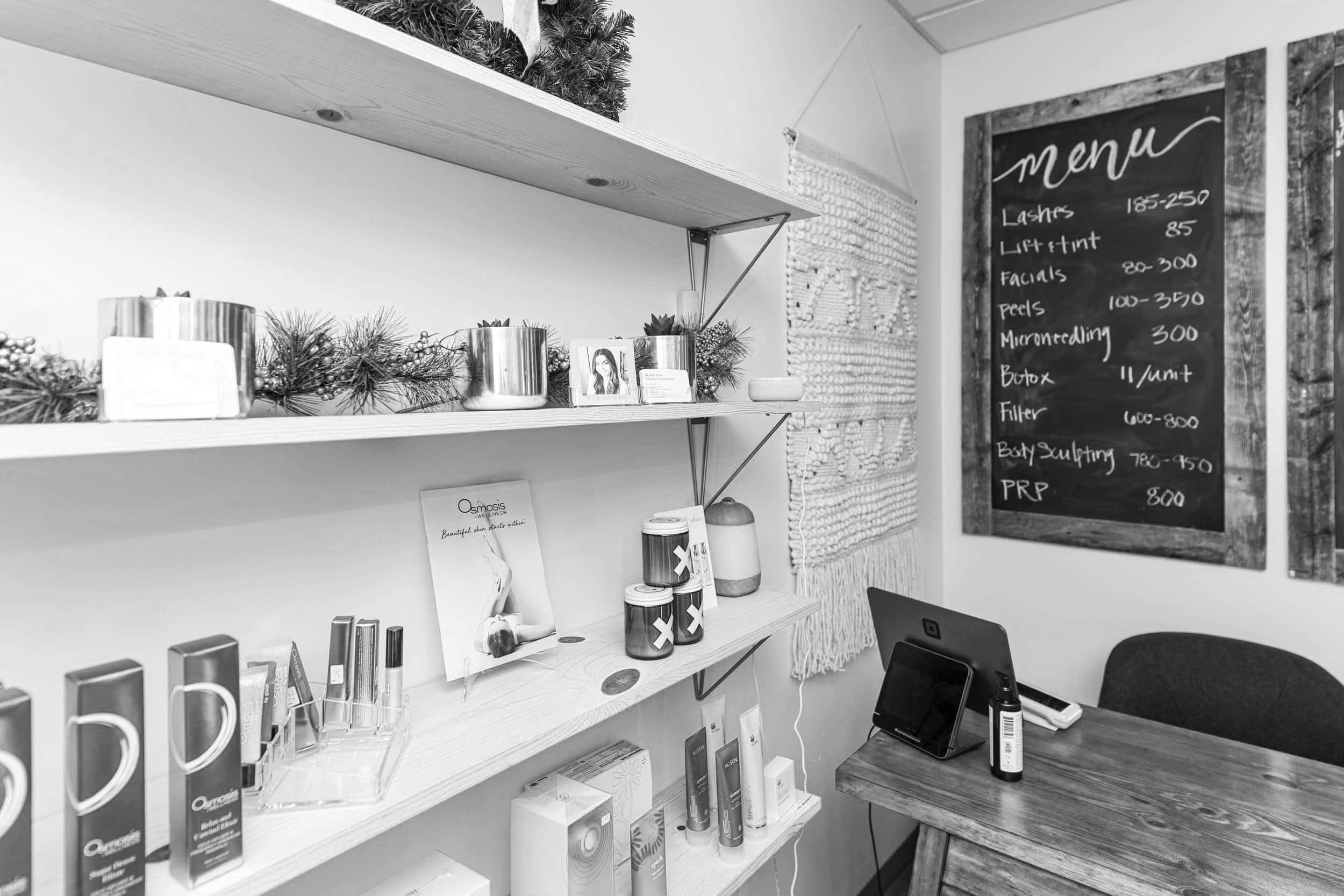 Black and white photo of a retail or medical setting with shelves displaying products, a chalkboard menu listing services and prices, and a desk with a laptop and some bottles.