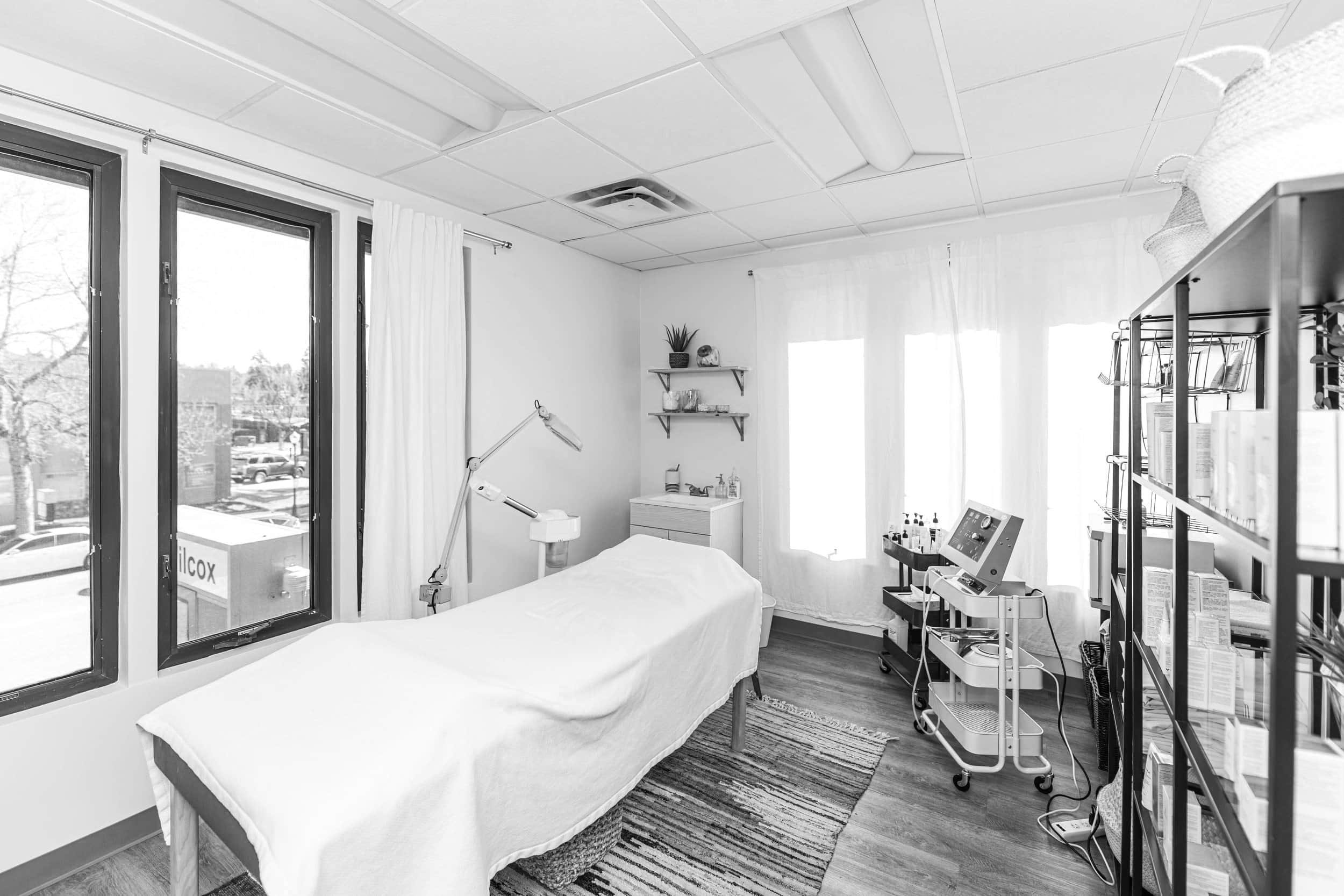 A spa treatment room with medical equipment, and shelves with supplies, illuminated by natural light through large windows.