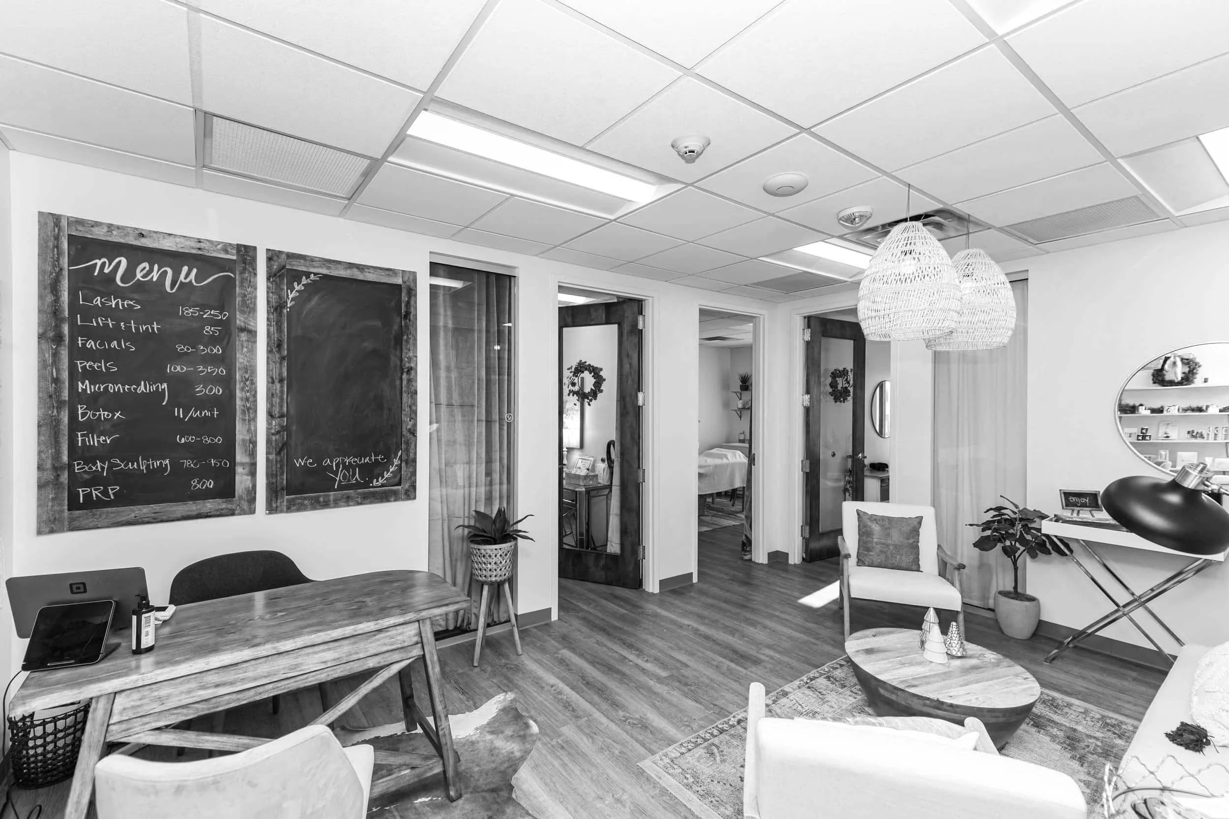 Interior view of a modern office or waiting area with a wooden table, chairs, and decorative plants. There are two chalkboards with menu items and prices, wooden doors with wreaths, and woven hanging lights.
