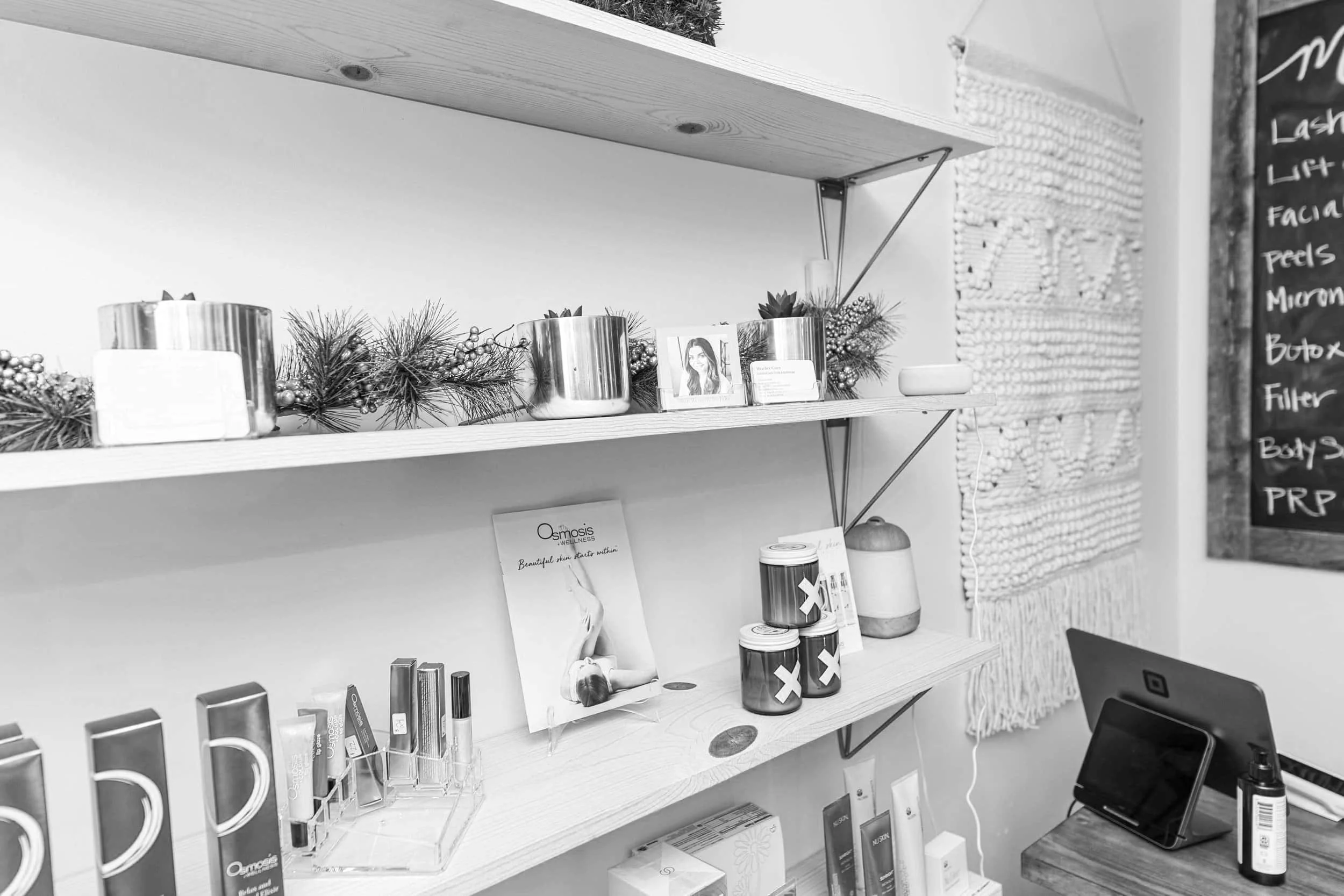 Black and white photo of a display shelf with skincare and wellness products, a business card holder with a photo of a woman, and a small potted plant. To the right, part of a chalkboard menu listing services such as lash lift, facials, and microblad