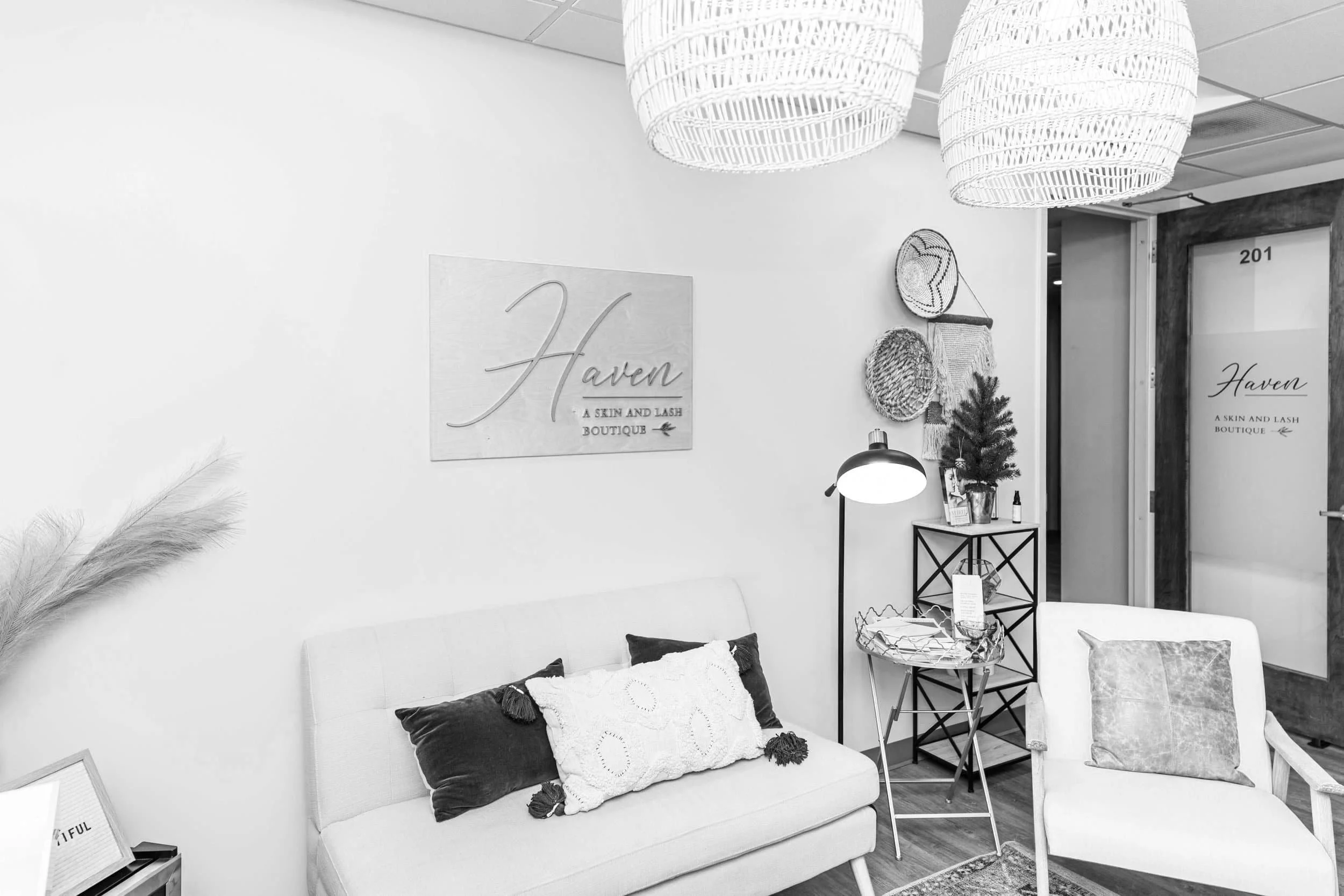 Interior of a boutique with a sofa adorned with pillows, a small table, wall decor including a sign that reads "Haven - A Skin and Lash Boutique," and hanging wicker light fixtures.