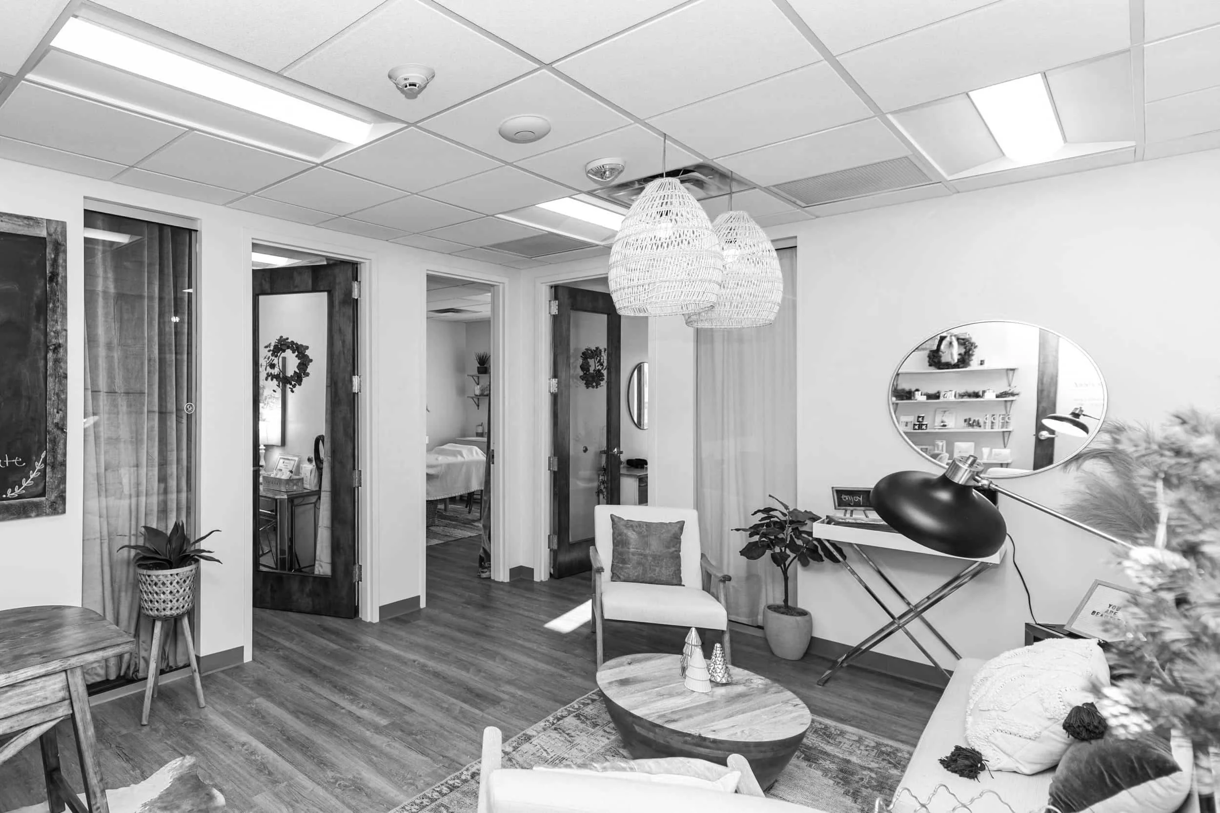 Cozy interior of a waiting or living area with wooden flooring, ceiling lights, hanging woven lamps, a round mirror, chairs, potted plants, and decorative Christmas items.