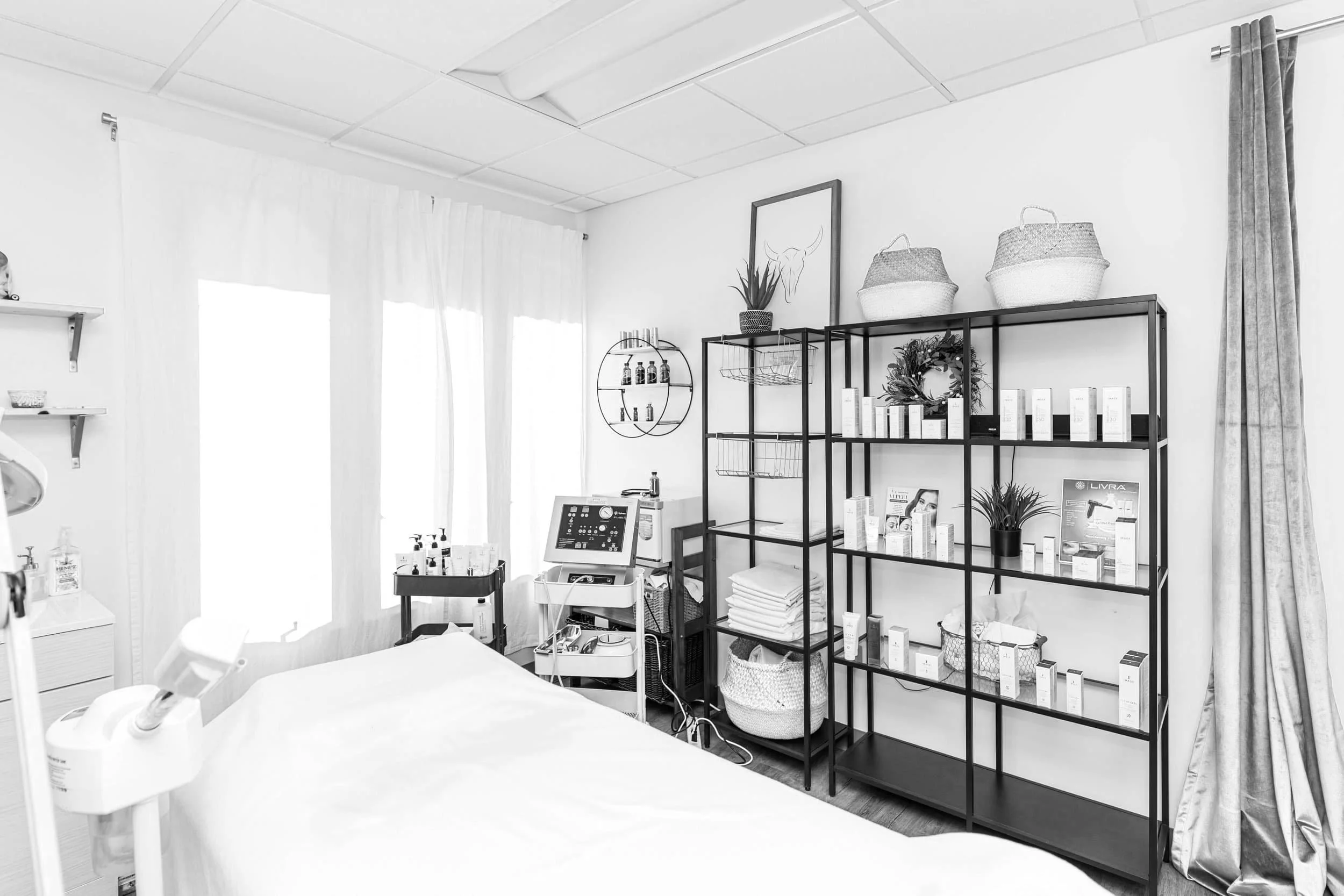 Medical examination room with a bed, shelves, and medical equipment, including bottles, boxes, and a monitor. Curtains cover windows, with decorative baskets and a framed artwork of a bull's head on the wall.