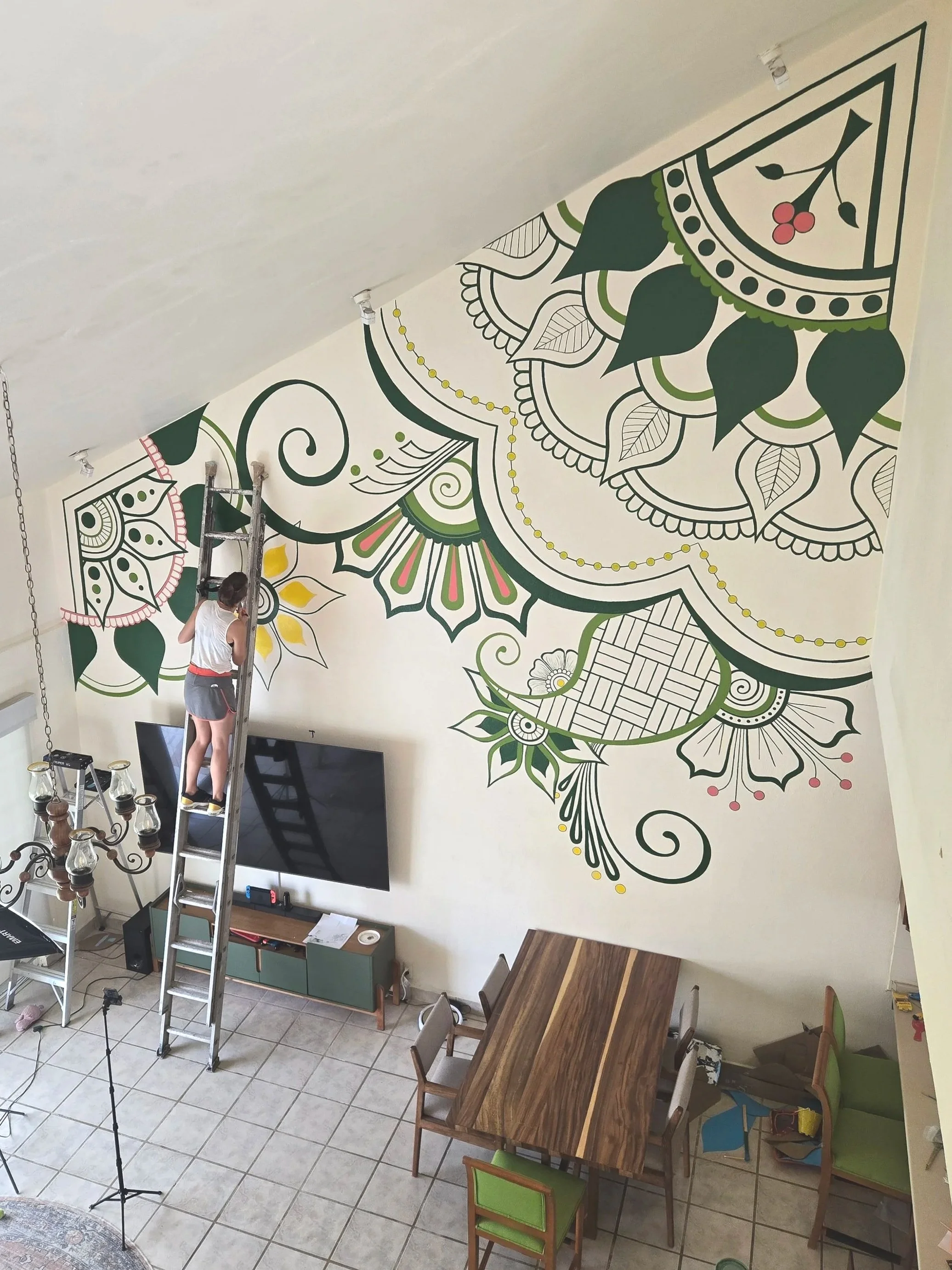 April Couvillion standing on a ladder painting a large custom Mural. An intricate yellow, green, and pink henna style design adds interest and vitality to this living space