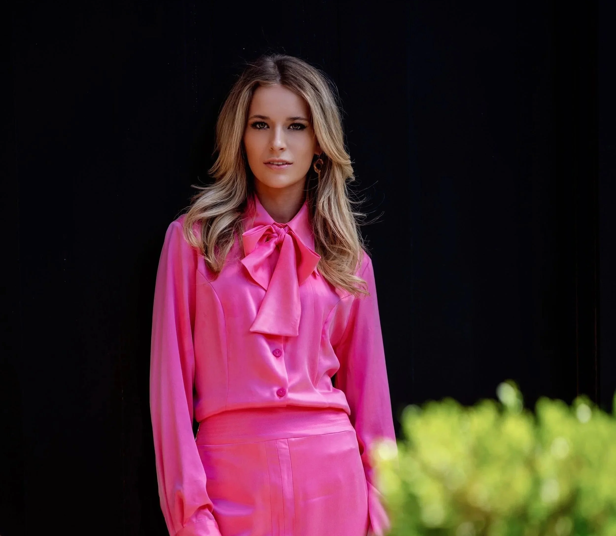 A woman with long wavy blonde hair wearing a bright pink satin blouse with a large bow and a matching pink skirt, standing outdoors against a black background with some greenery in the foreground.