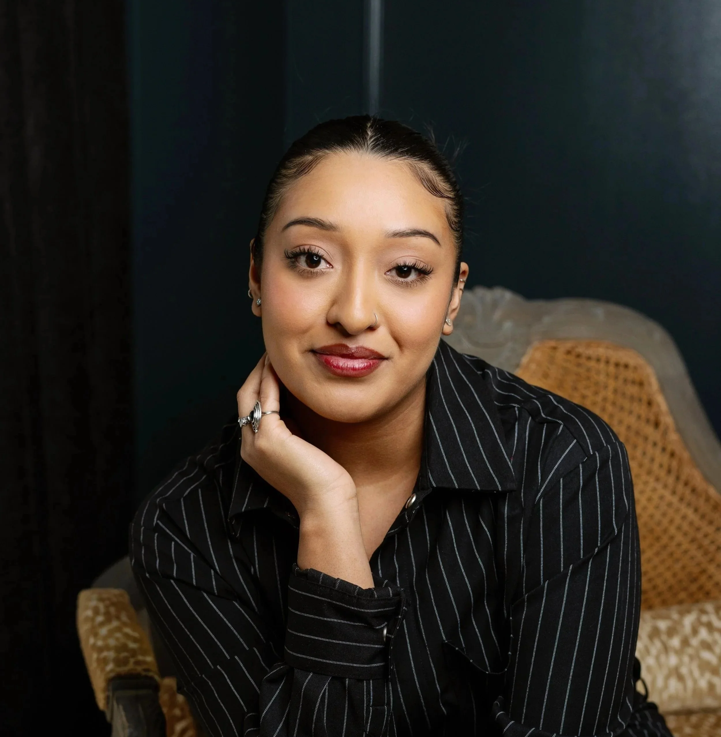 A young woman with dark hair tied back, wearing a black pinstripe shirt, sitting on a wicker chair with a dark background. She has a gentle smile and touches her face with her hand.