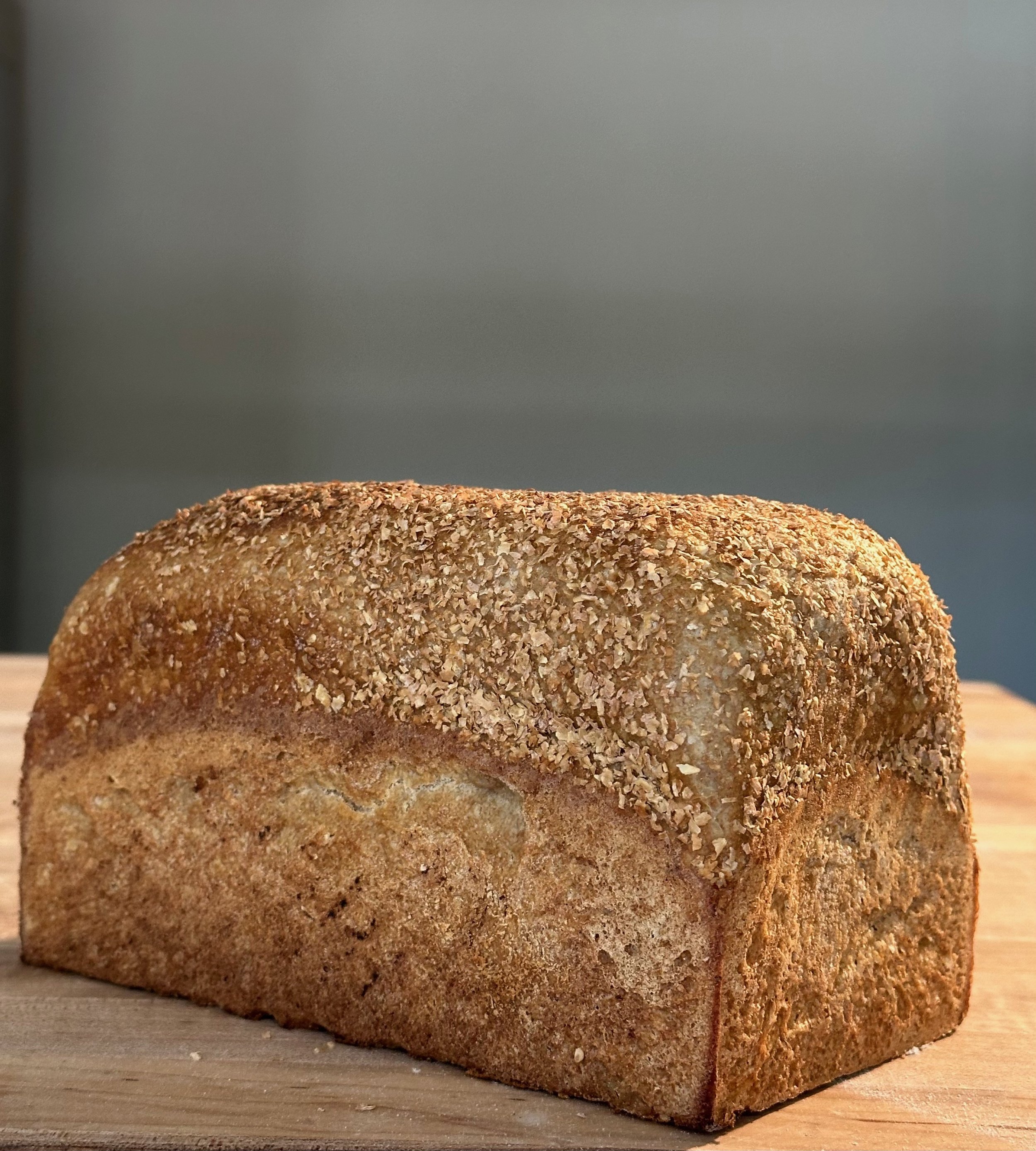 A loaf of bread with a crispy, golden-brown crust topped with wheat bran , resting on a wooden table.