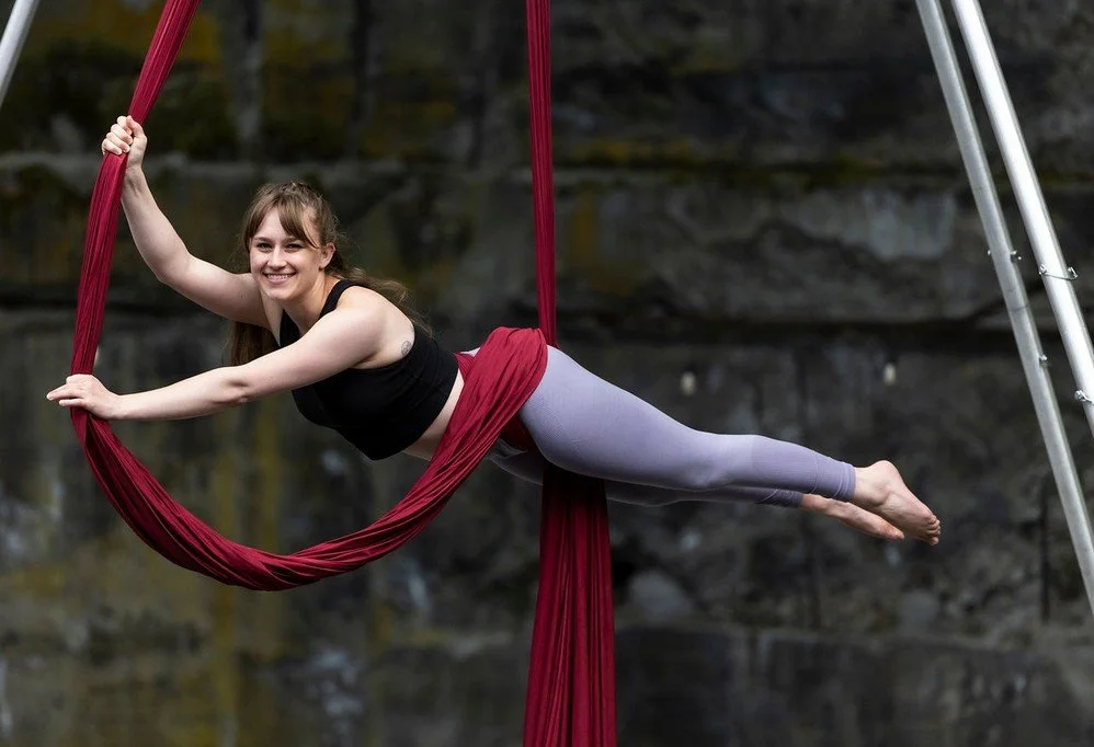 Children practicing aerial silk gymnastics indoors, hanging upside down and smiling, with colorful silks and a padded floor.