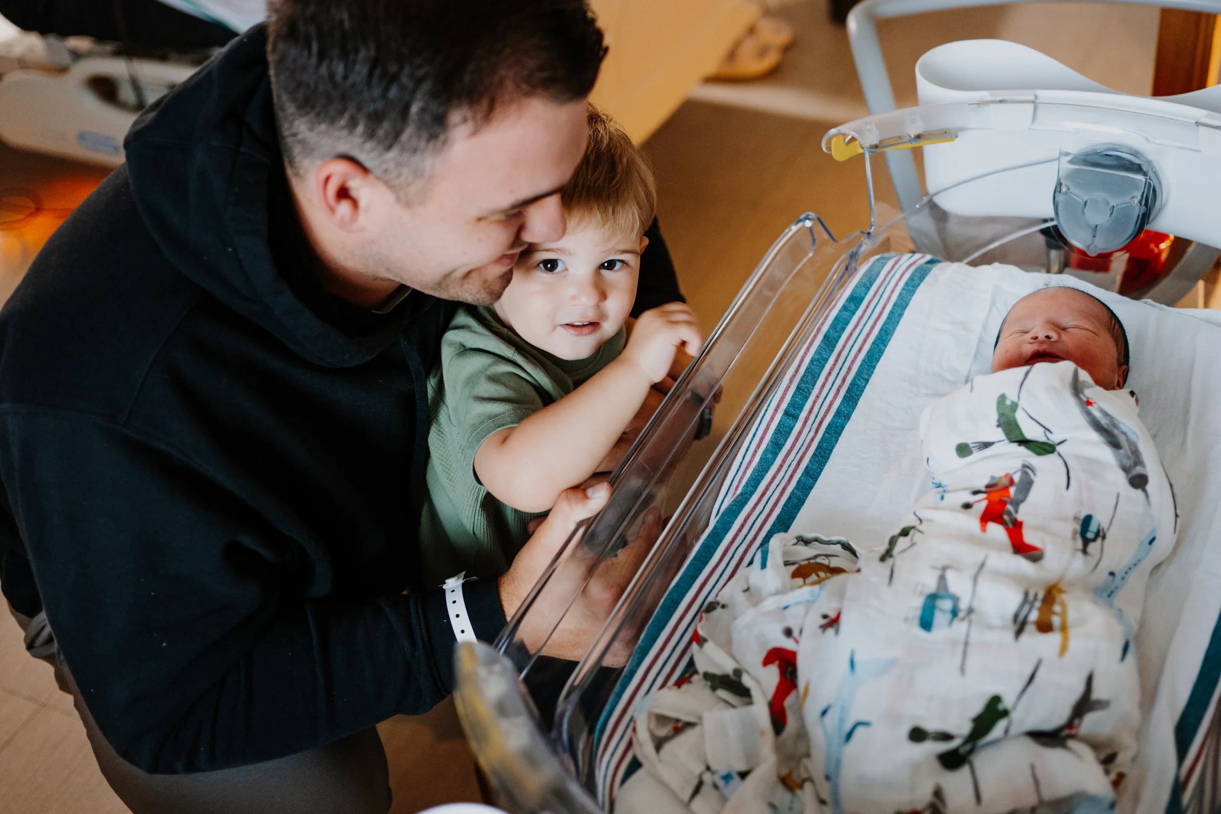 A man and a young boy are observing a newborn baby in a bassinet at a hospital. The boy looks curious, and the man is smiling, both leaning close to the baby.