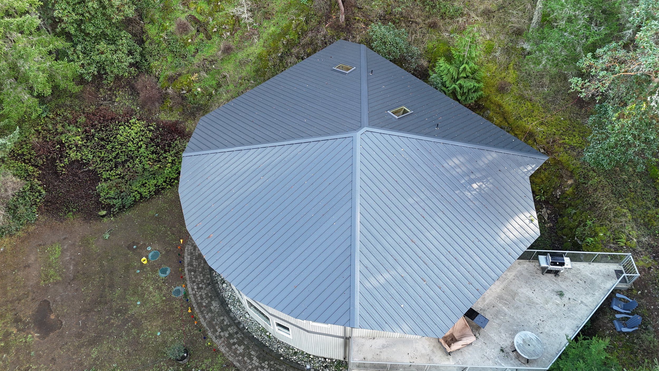 Aerial view of a house with a gray metal roof surrounded by green trees and a backyard with a concrete deck, barbecue grill, and outdoor chairs.
