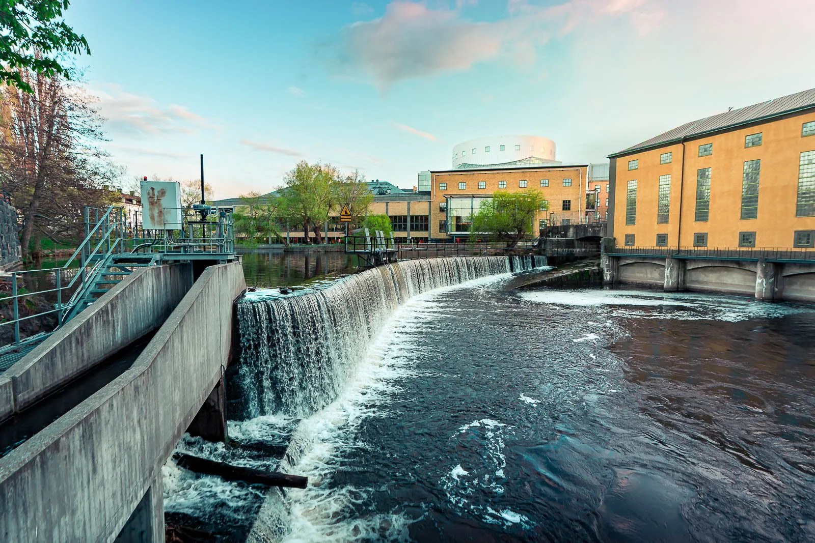 En stadssjö med ett fallande vattenfall framför gula och gula byggnader, träd och en blå himmel.