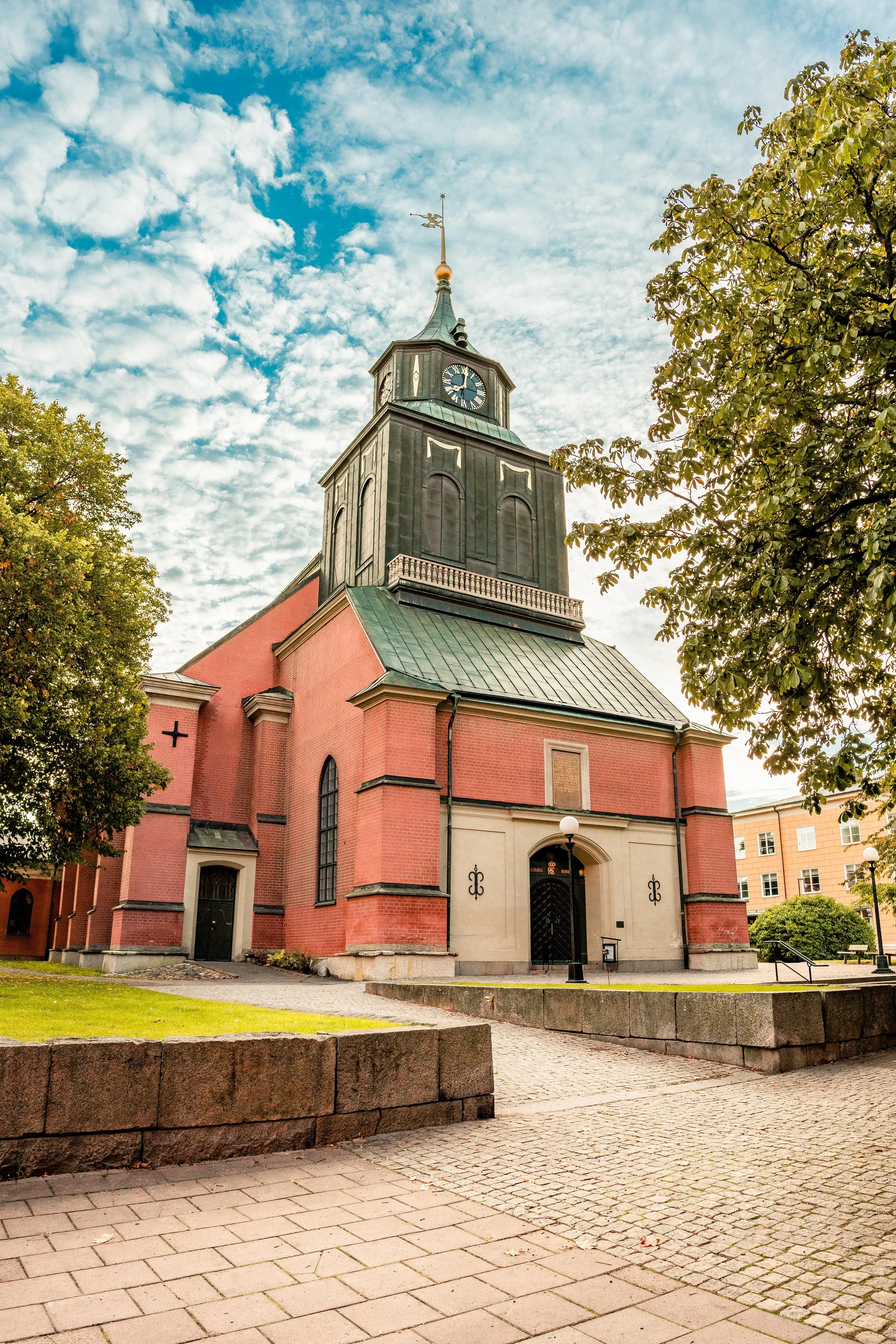 Foto av en kyrka med en hög spira och en klocka, omgiven av träd och en stenmur, under en molnig himmel.