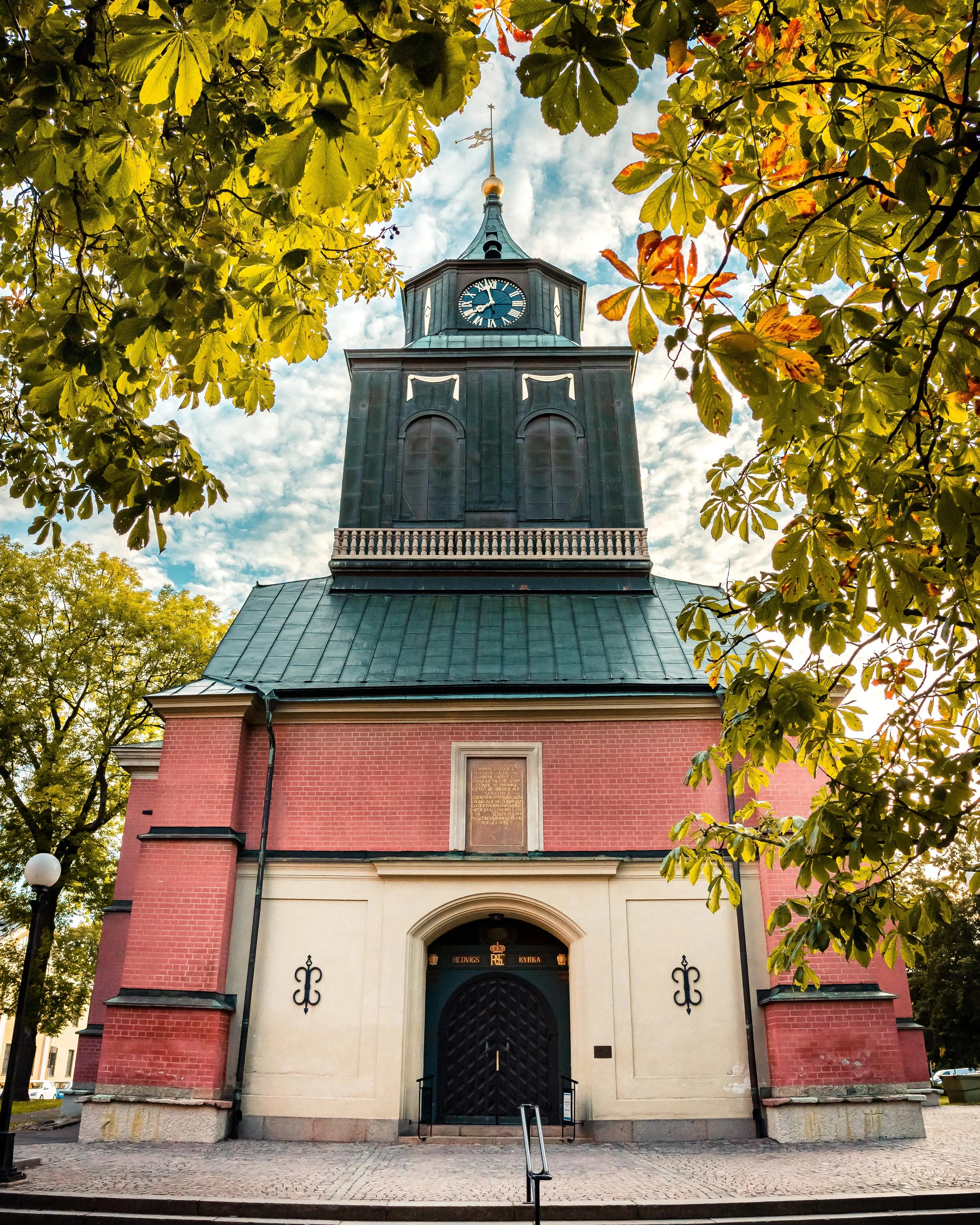 Bild av en kyrka med en mörkgrön tornspira och en klocka, omgiven av träd med gröna och gula blad, tydligt under en dag med molnigt väder.