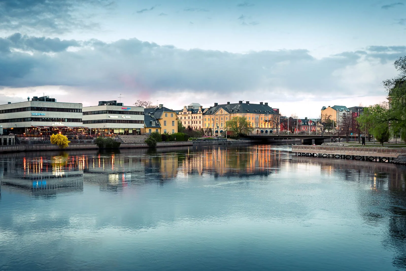 Stadsbild med en flod, moderna och äldre byggnader, samt en bro under en molnig himmel