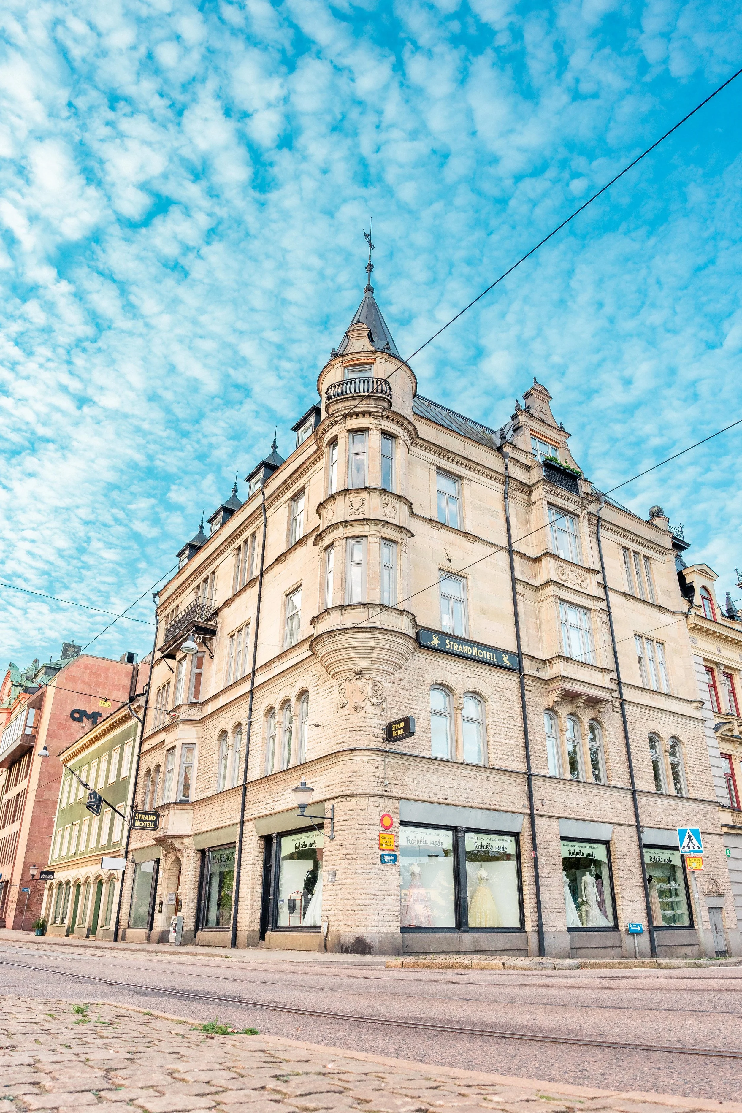 Ett gammalt, flera våningar högt hotell med en tornliknande utbyggnad, skylt som säger 'Strand Hotell', stora skyltfönster på bottenvåningen med klädbutiker, tram- eller tågspår på gatan framför byggnaden, och en blå himmel med moln.