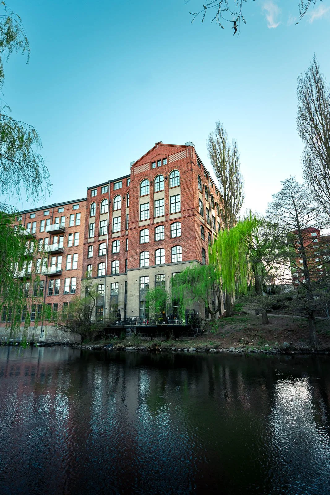 Arkitekturbild från Norrköping, fotograferad av BKMEDIA, Badi Khwies. Bilden visar en röd tegelbyggnad vid Motala ström med träd, vatten och speglingar i förgrunden. Visuellt innehåll för platsmarknadsföring och destinationer