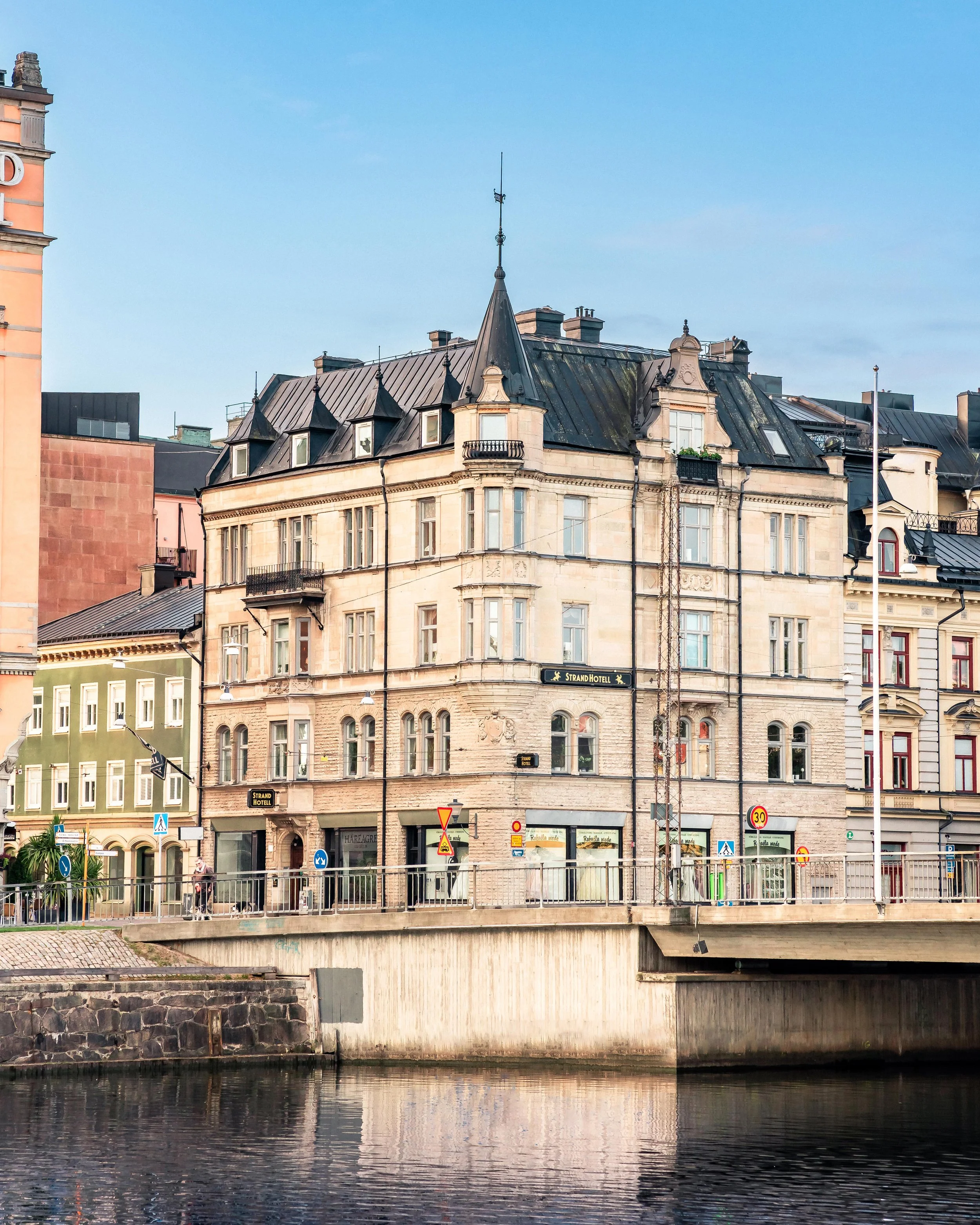Historisk byggnad vid en kanal i en stad med bilar och människor som promenerar på gatan framför. Solig dag med blå himmel.