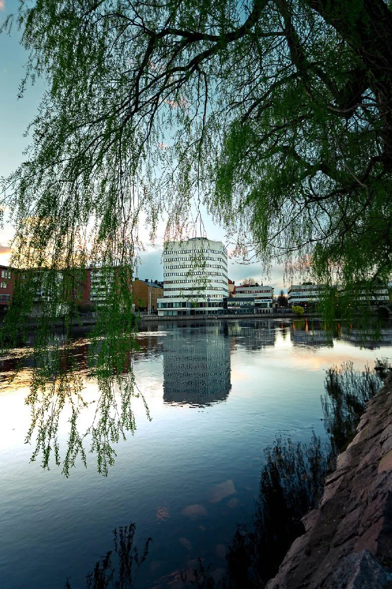 Stadsbild från Norrköping, fotograferad av BKMEDIA, Badi Khwies. Bilden visar Holmenområdet speglat i Motala ström, inramat av grenar och kvällsljus. Visuellt innehåll för platsmarknadsföring, destinationer och stadsmiljöer.