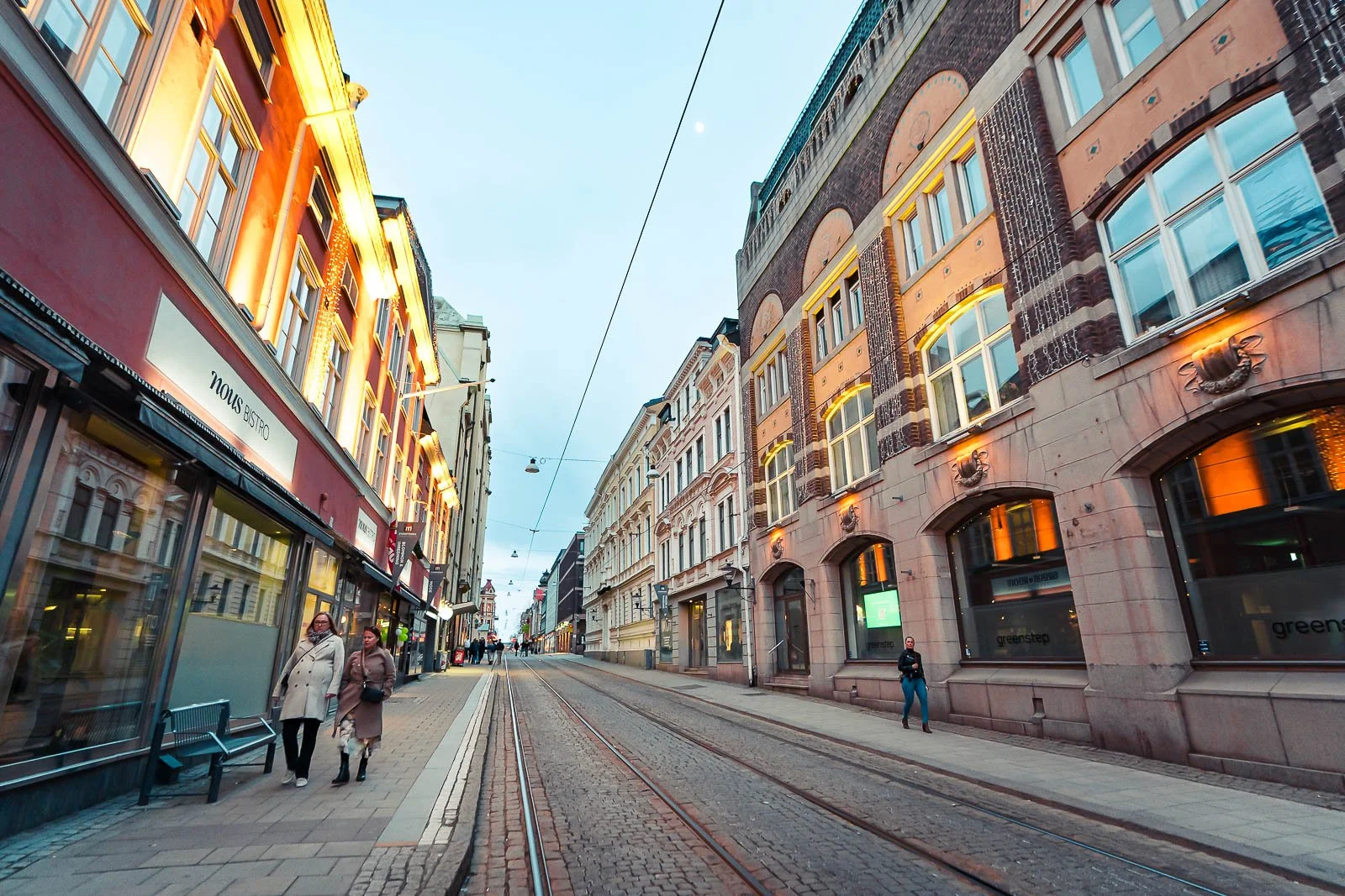 En stadsgata med gamla byggnader och spårvagnsskenor i mitten, under kvällstid, med människor som går på trottoaren.