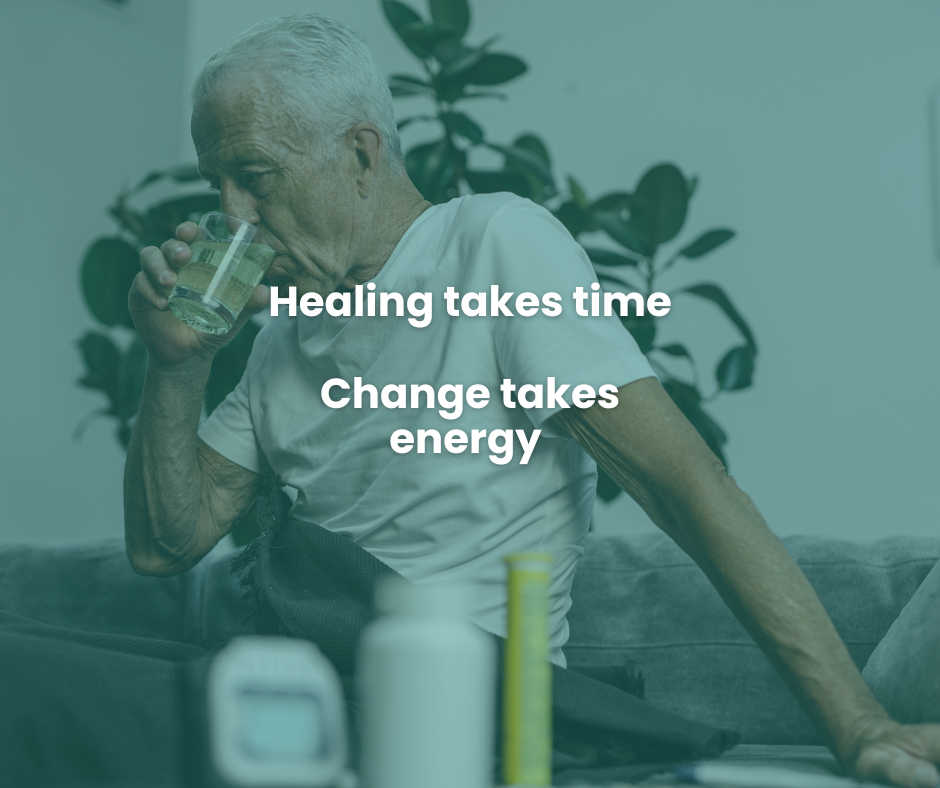 An elderly man drinks water while sitting indoors with a potted plant in the background, surrounded by bottles and supplements.