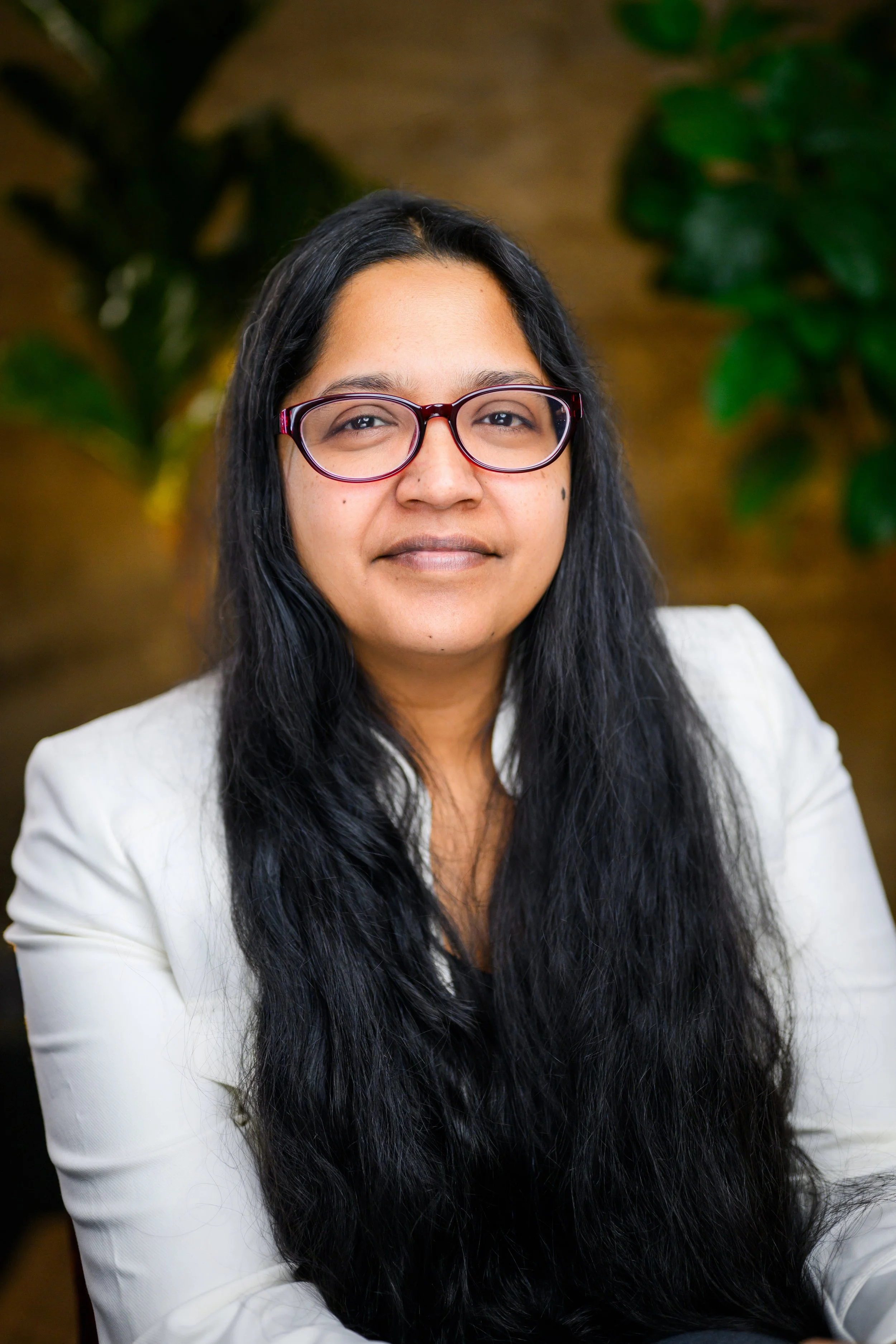 A woman with long black hair, glasses with reddish frames, wearing a white blazer, sitting in front of a wooden background with green plants.
