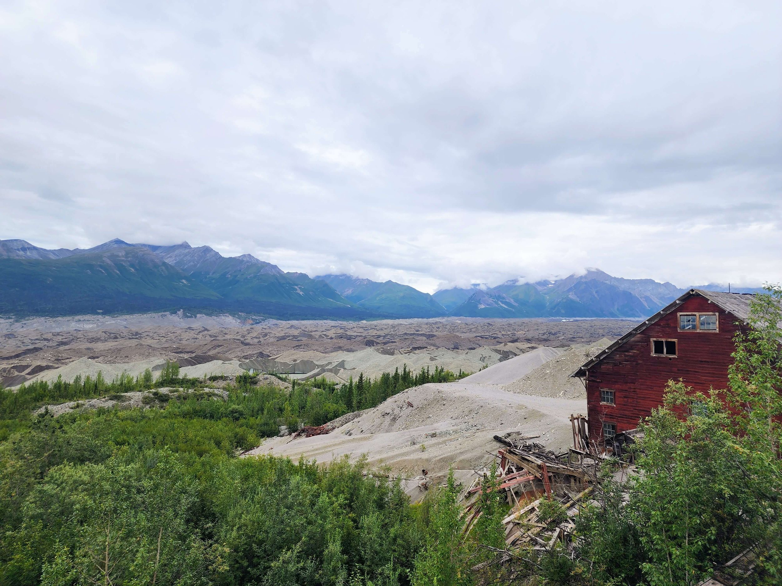 View of Root Glacier covered in silt, so much so that it looks like miles of dirt, with the remains of a red wooden mill building in the foreground.