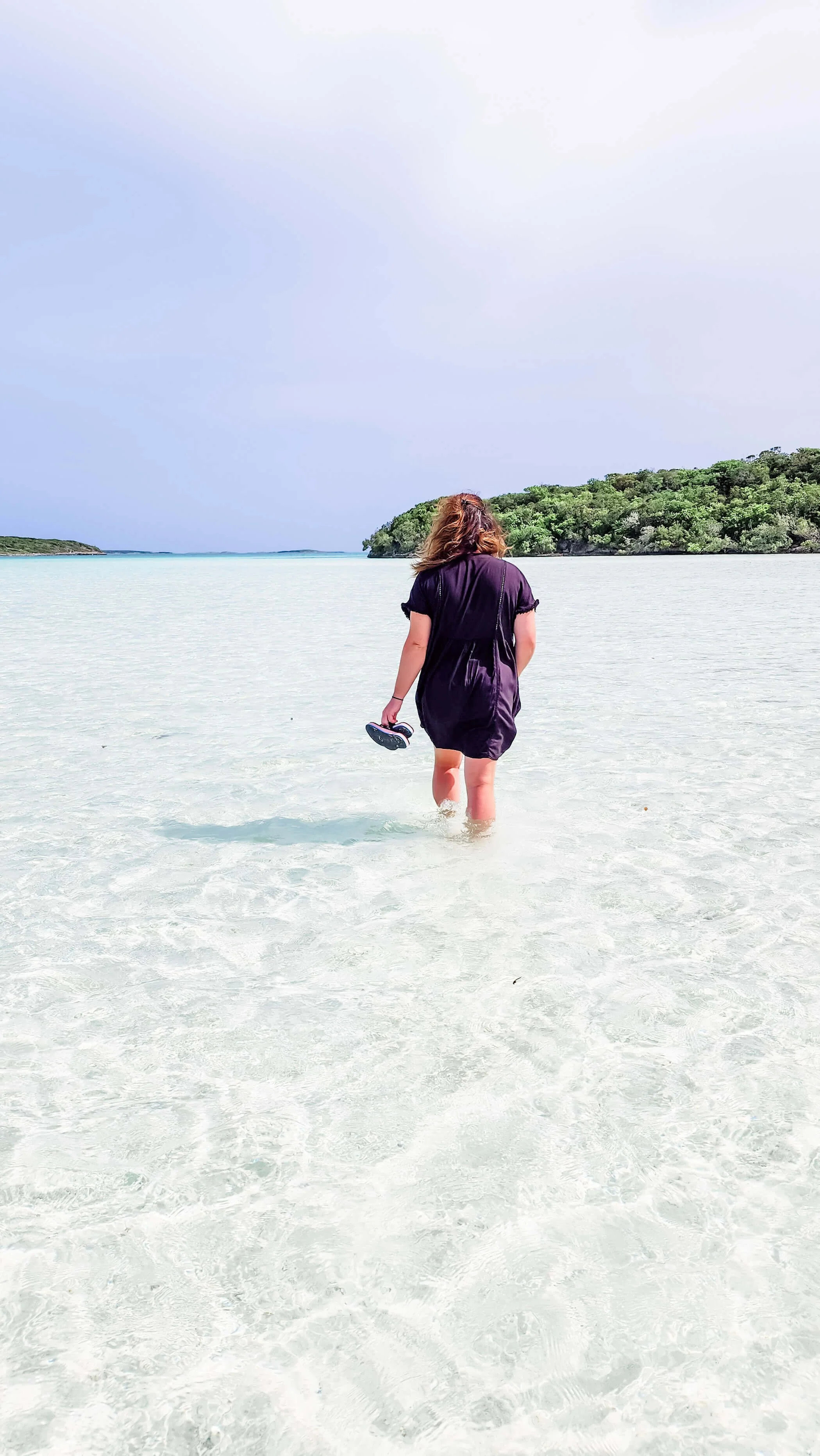 Girl wearing a black swimsuit coverup walking in shallow, clear, blue water at Exuma Point in the Bahamas.