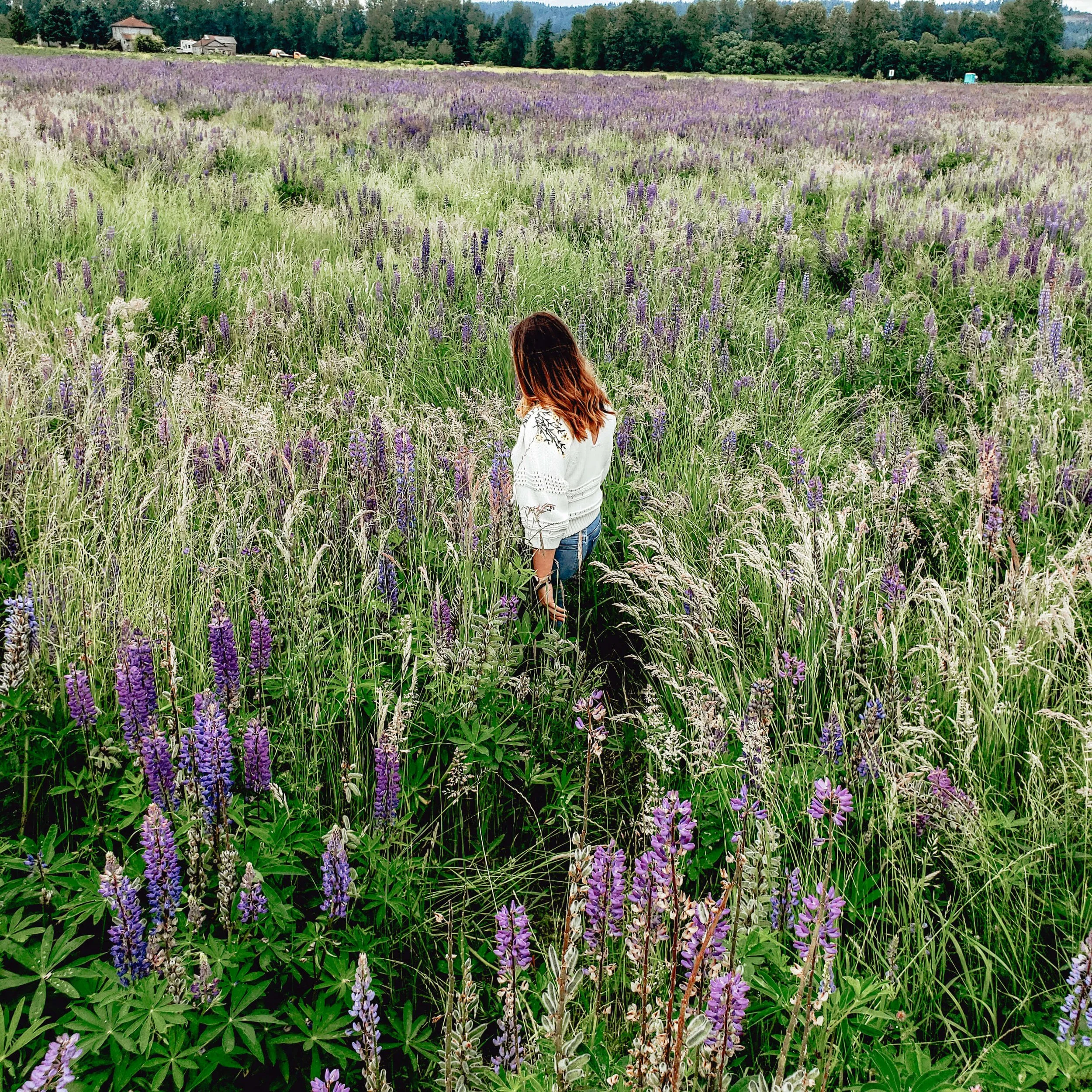 Young woman walking through a field of tall grass and purple flowers, surrounded by a countryside landscape.