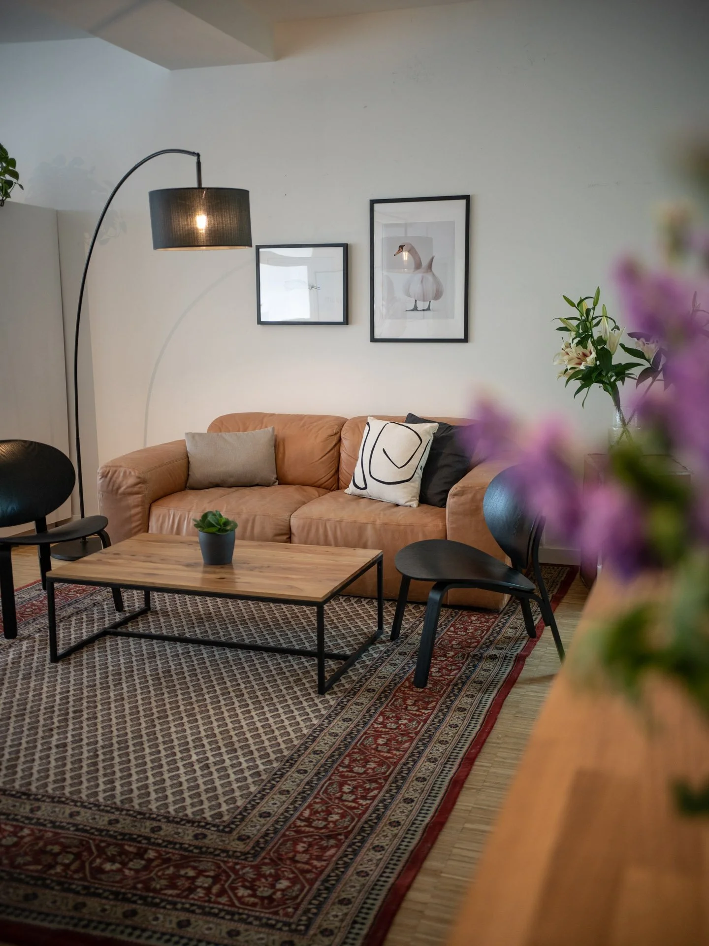 Produktionsküche | Mietküche in Köln | Gemütliches Wohnzimmer mit braunem Ledersofa, Kaffetisch, schwarzen Stühlen, Stehlampe, Bildern an der Wand und Blumen auf einem Regal.