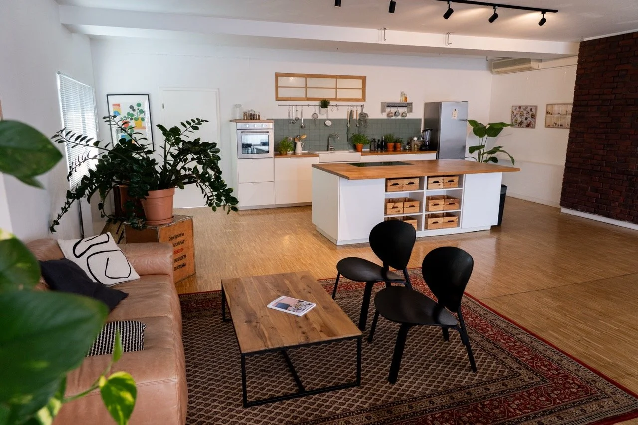 Offenes Wohnzimmer und Küche mit Holzmöbeln, schwarzen Stühlen, grünen Pflanzen, dekorativen Wandbildern und einem roten Teppich.