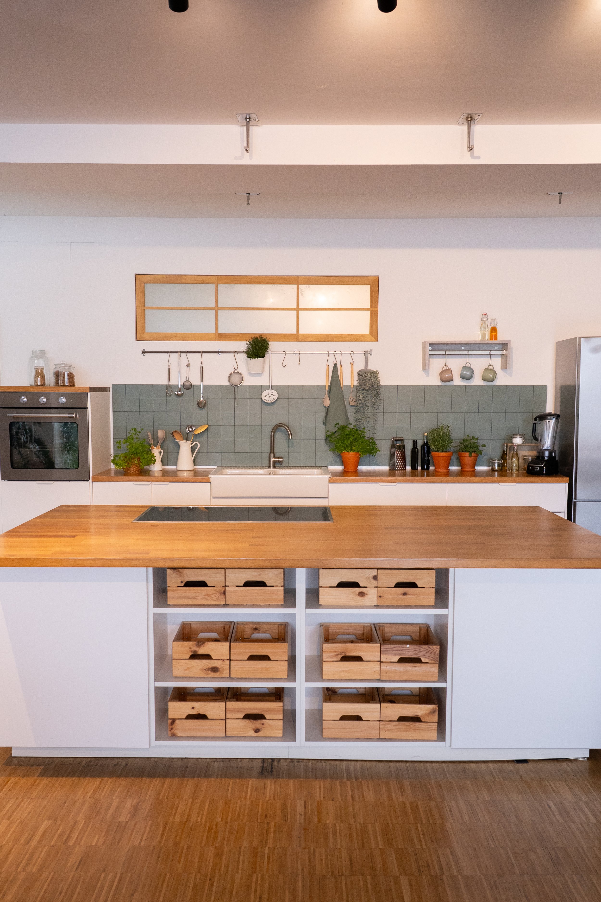 Moderne Küche mit Holzarbeitsplatte, grüner Fliesenspiegel, Pflanzen in Töpfen, Küchenutensilien, Backofen, Kühlschrank und aufgeräumten Schubladen und Regalen.