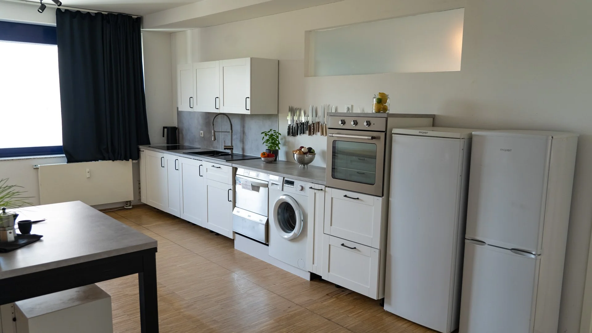 Produktionsküche | Vorbereitungsküche | Blick auf eine moderne weiße Küchenzeile mit Eingangstür, Geschirr, Herd, Waschmaschine und Kühlschrank in einer hellen Wohnung.