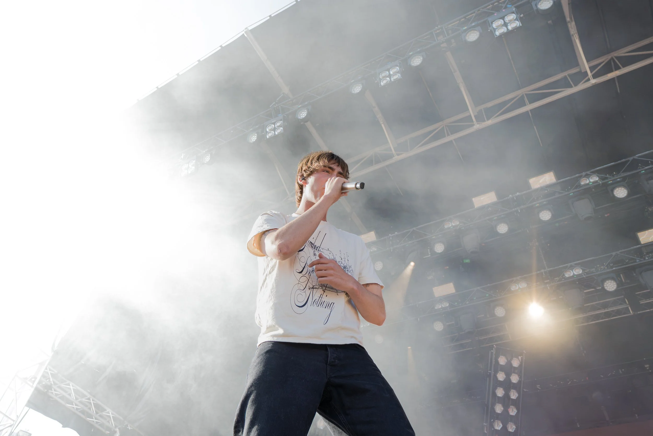 En ung mand med mikrofon i hånden, optræder på en stor scene med lysudstyr og røg, ved en koncert eller festival.