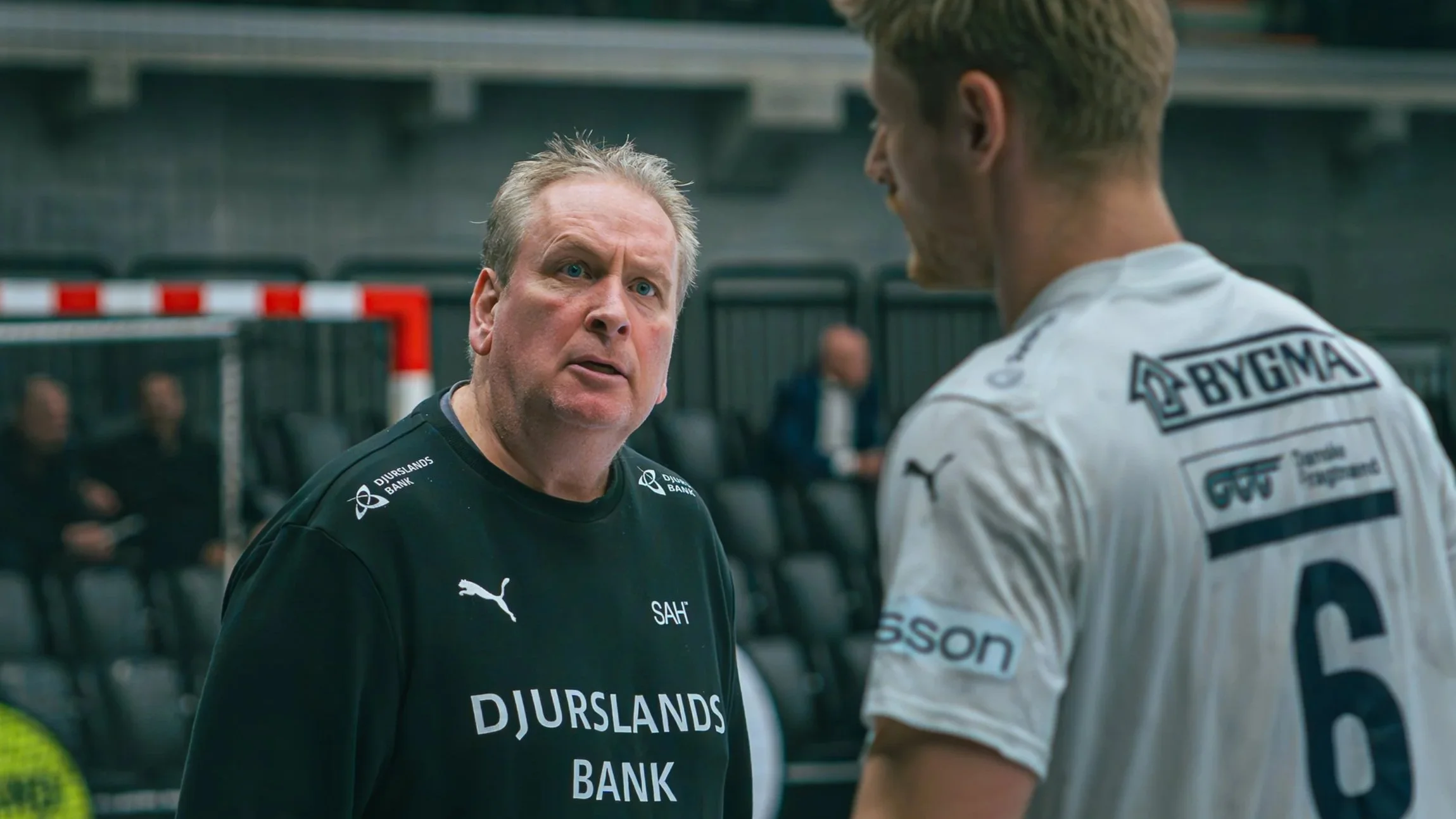 To mænd i en sportsarena, den ene i sort trøje med teksten 'DJURSLANDS BANK', den anden i hvid trøje med sponsorlogoer.