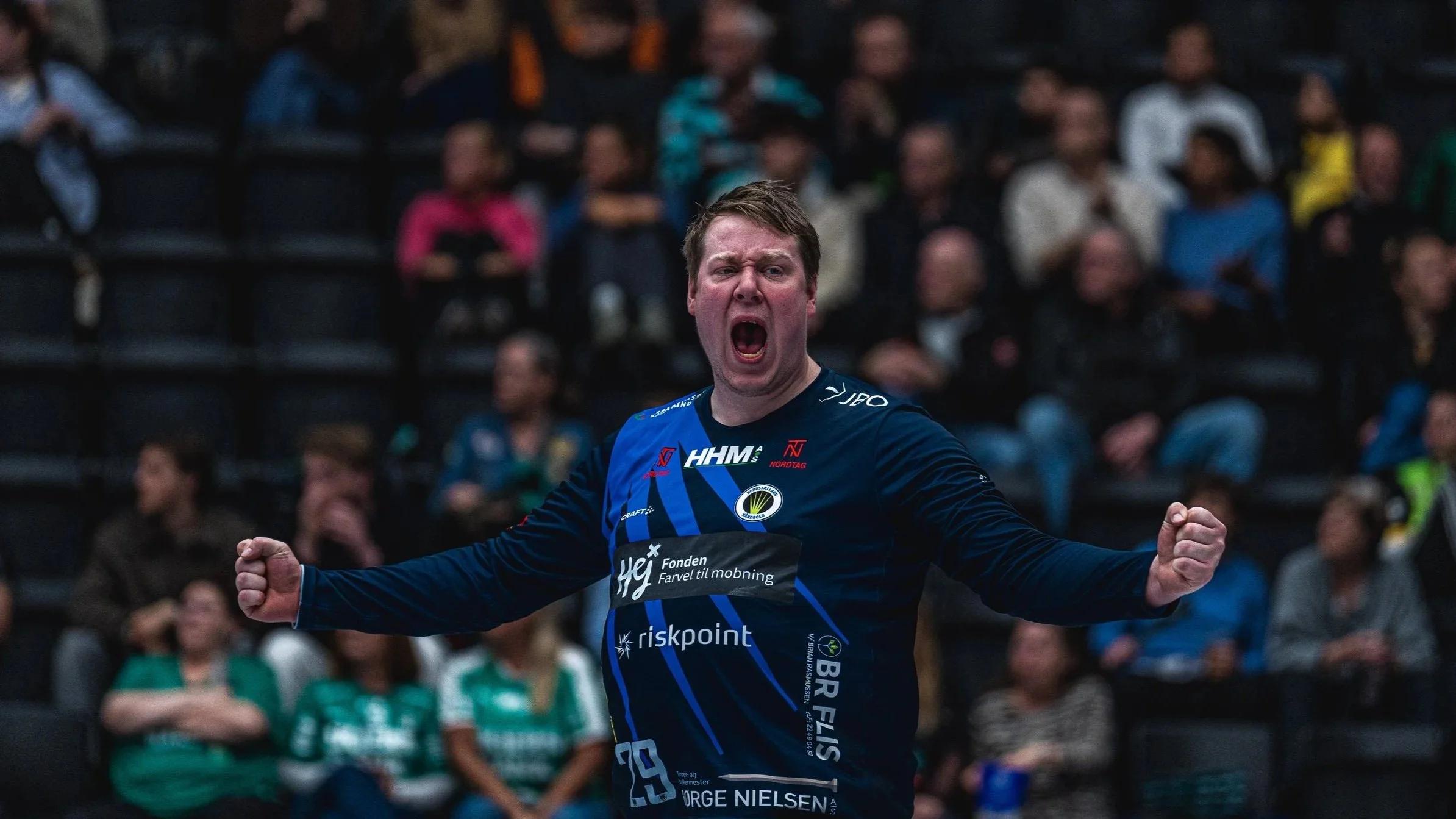En mand i en blå sporttrøje udtrykker glæde og triumf med hænder i luften, i en indendørs sportsarena med tilskuere i baggrunden.