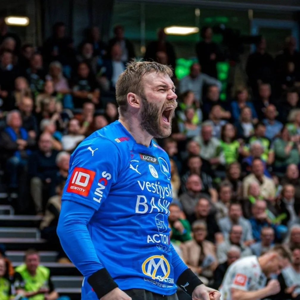 En mand i en blå sporttrøje jubler med knyttede næver under en håndboldkamp, omgivet af tilskuere i en sportsarena.