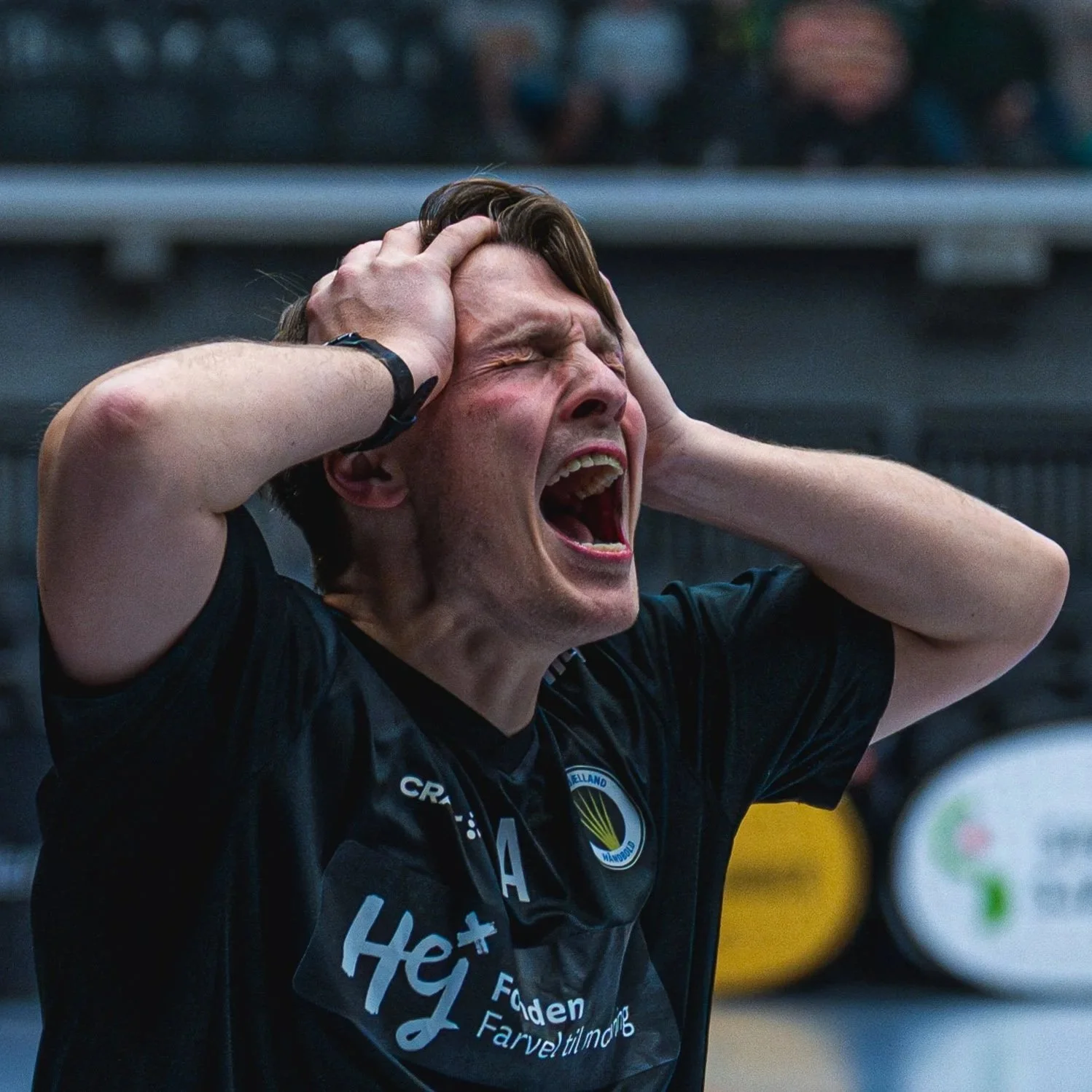 En mand med kort hår, iført en sort sports T-shirt, holder sin hoved med begge hænder med et tydeligt udtryk af frustration eller smerte, foran en sportsarena med tilskuere i baggrunden.