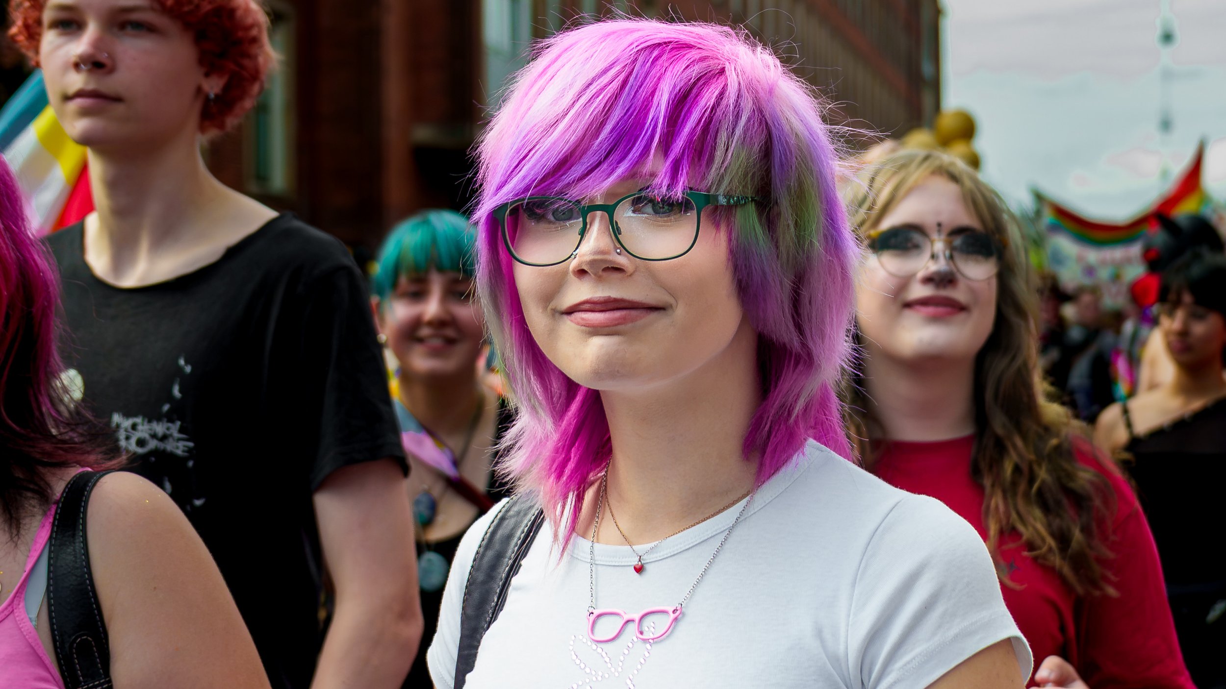 En ung kvinde med pink og grønt hår, iført store briller og en hvid t-shirt, står blandt en gruppe mennesker, der deltager i en festival eller et udendørs arrangement.