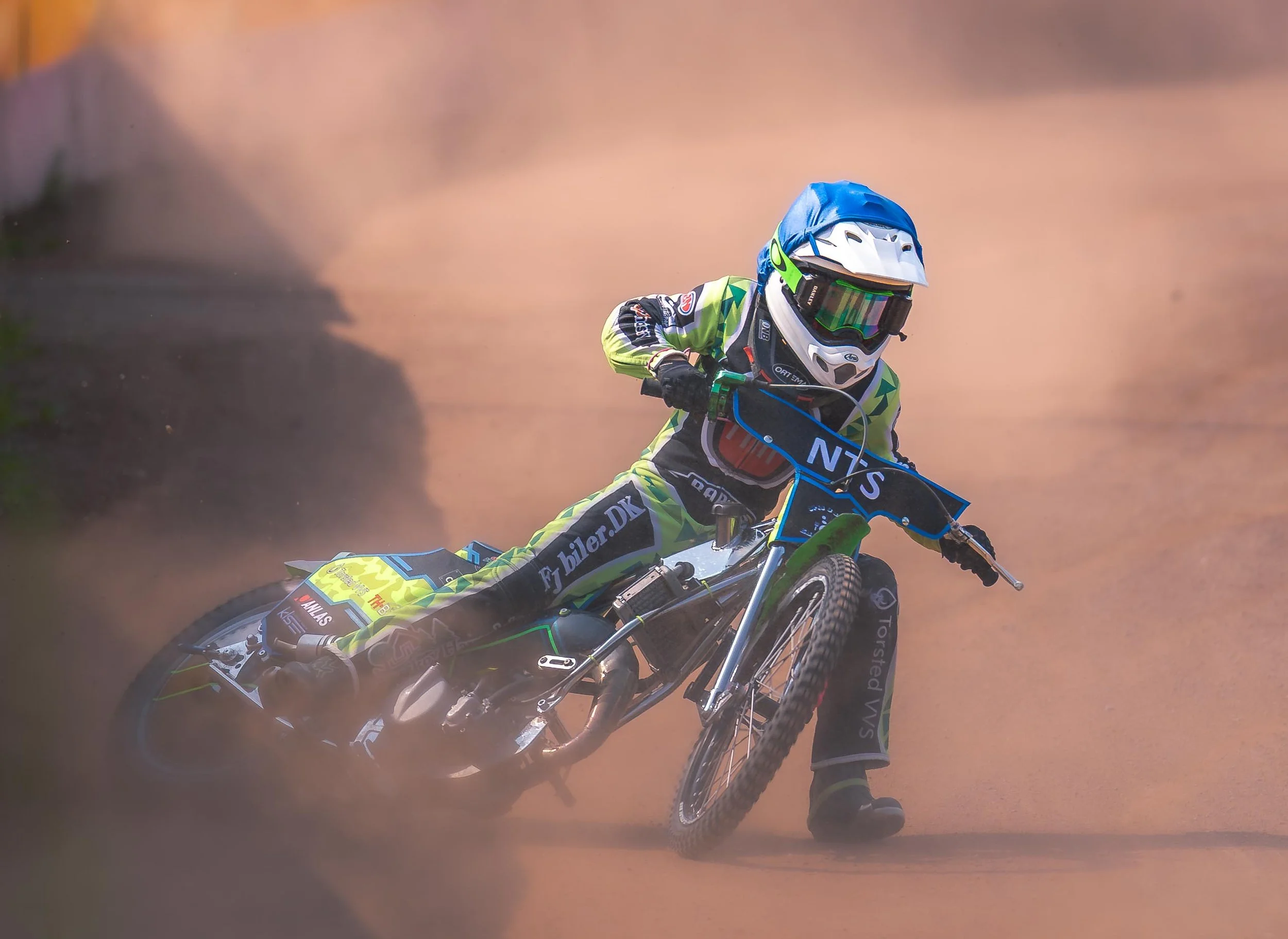 En motocrossfører i fuldt udstyr kører på en mudret bane, iført hjelm, beskyttelsesudstyr og farverigt tøj.