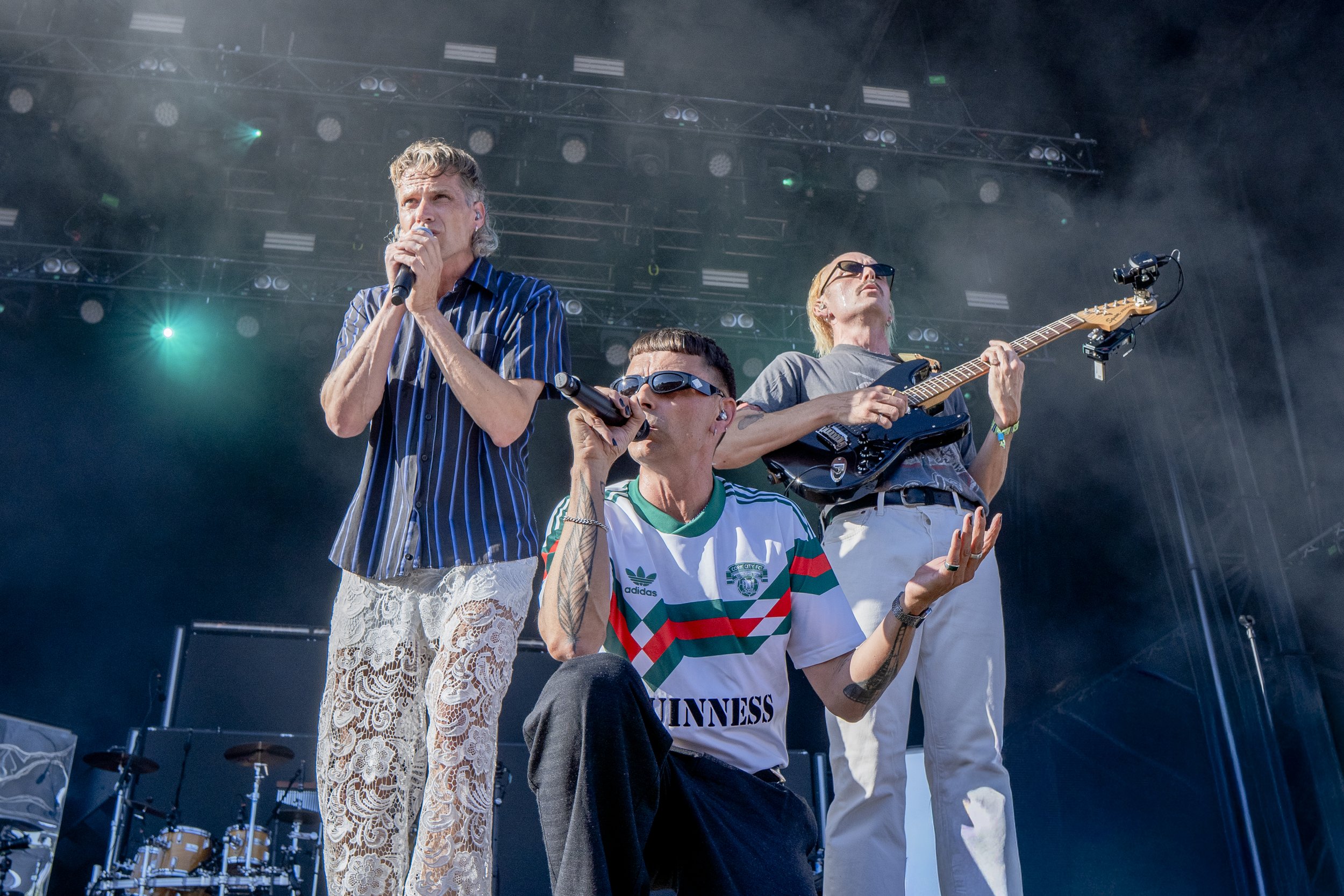 Fire mænd, der optræder på en scene ved en koncert. To af mændene synger i mikrofoner, mens den tredje spiller guitar. Baggrund med scenelys og trommer.