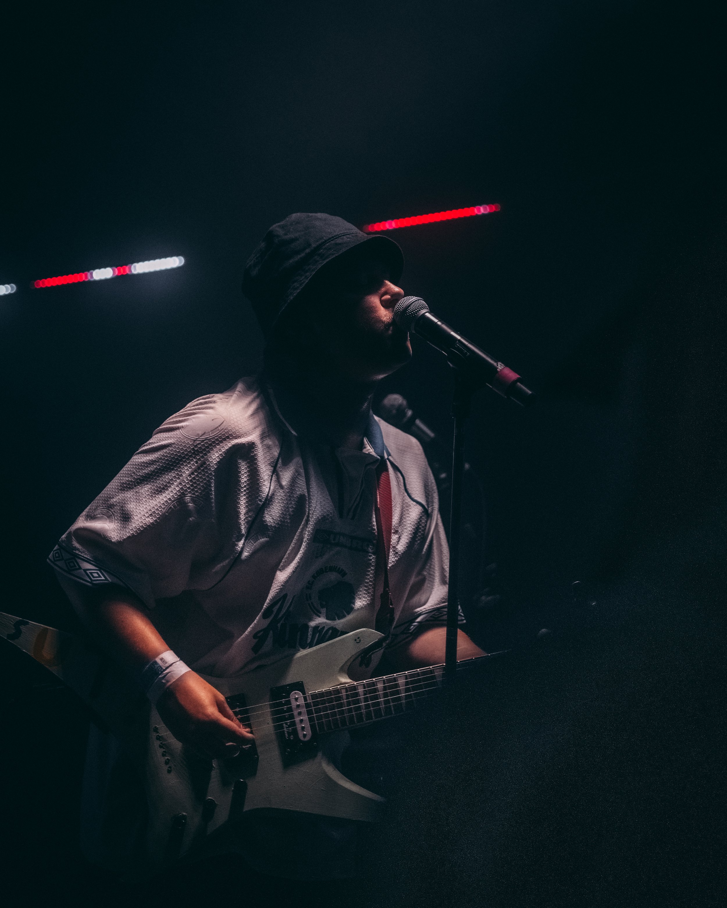 Person spiller guitar og synger ind i mikrofon i mørkt miljø, iført hoodie og sportstøj.