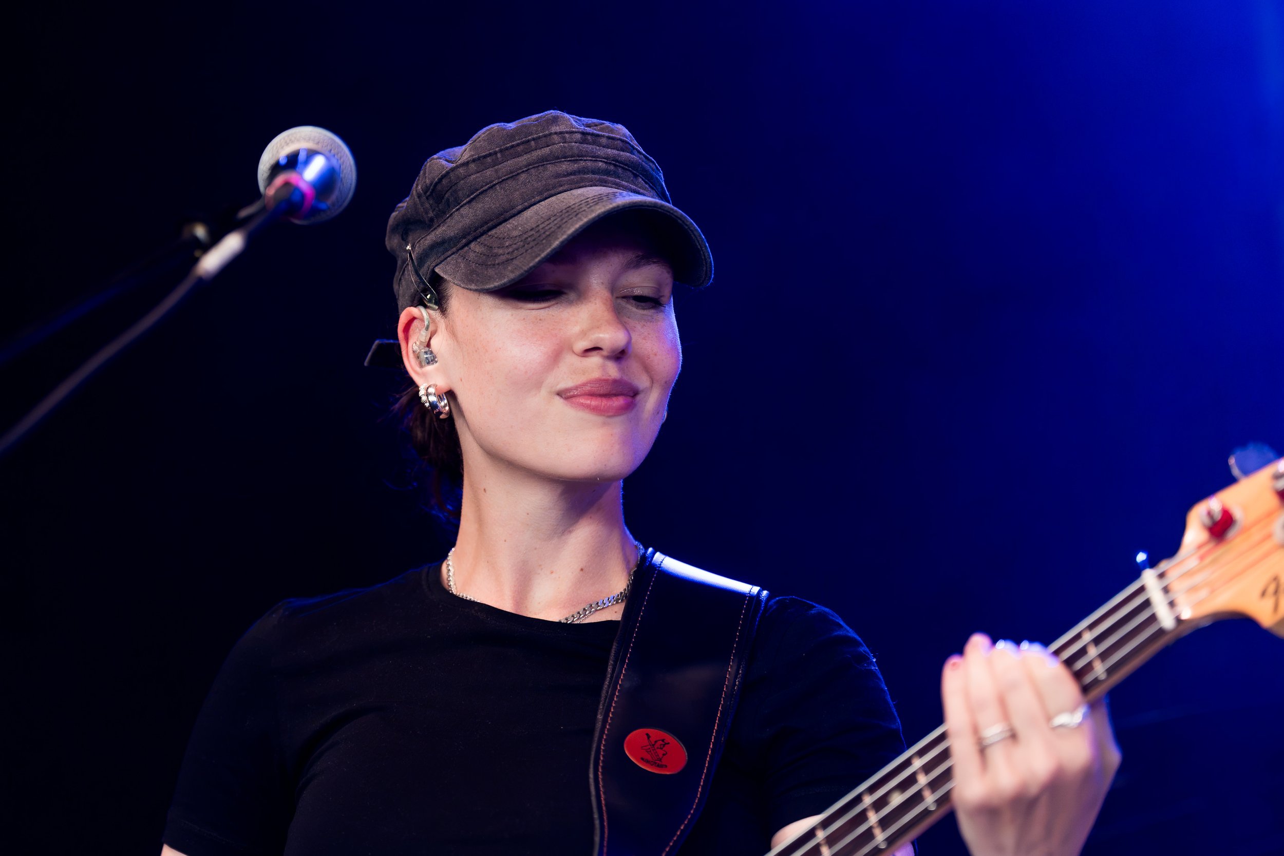 En woman med sort cap, smykker og sorte tøj spiller guitar på en scene med blå lys i baggrunden.