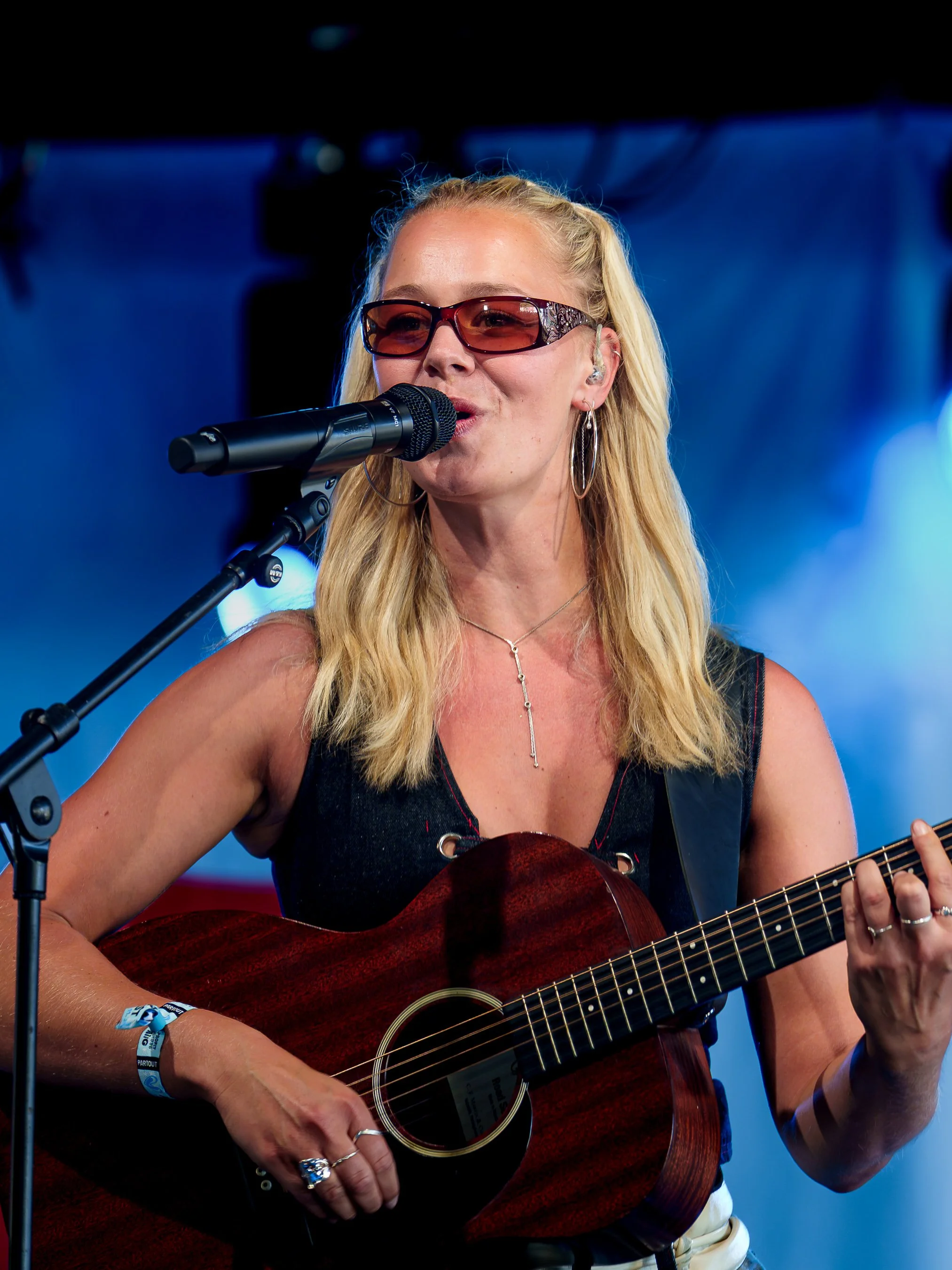 En kvinde med blond hår, solbriller, og store hoop øreringe spiller og synger i mikrofon og guitar på scenen, med blå baggrund og lys.