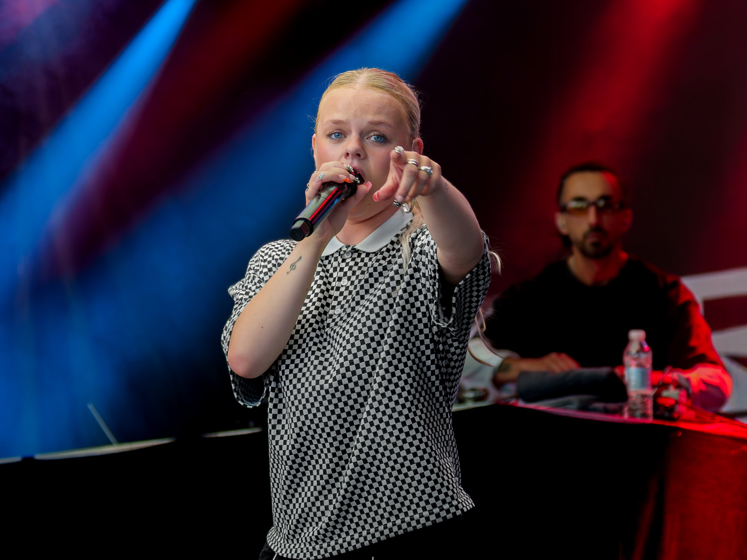 En kvinde med blondt hår og blå øjne holder mikrofon, peger mod kameraet, iført sort og hvid ternet skjorte. Bag hende, en mand med briller sidder bag DJ-udstyr. Farvet lys i rød og blå toner.