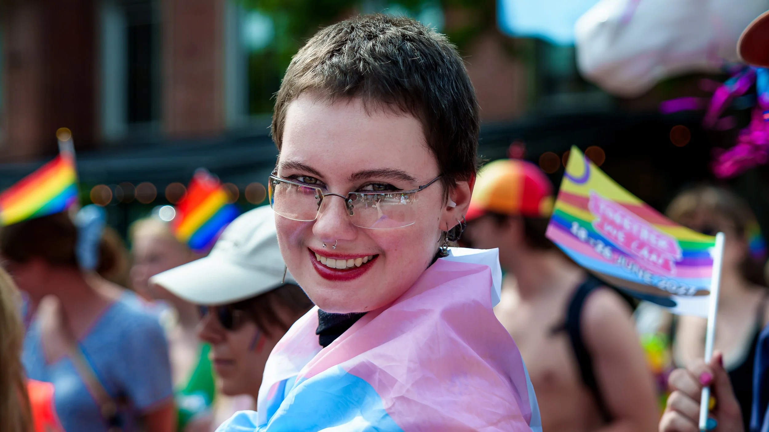 En person med kort mørkt hår, briller, og piercings i næse og ører smiler til kameraet under et pride-arrangement, omgivet af glade mennesker iført farverige t-shirts og pride-flags.
