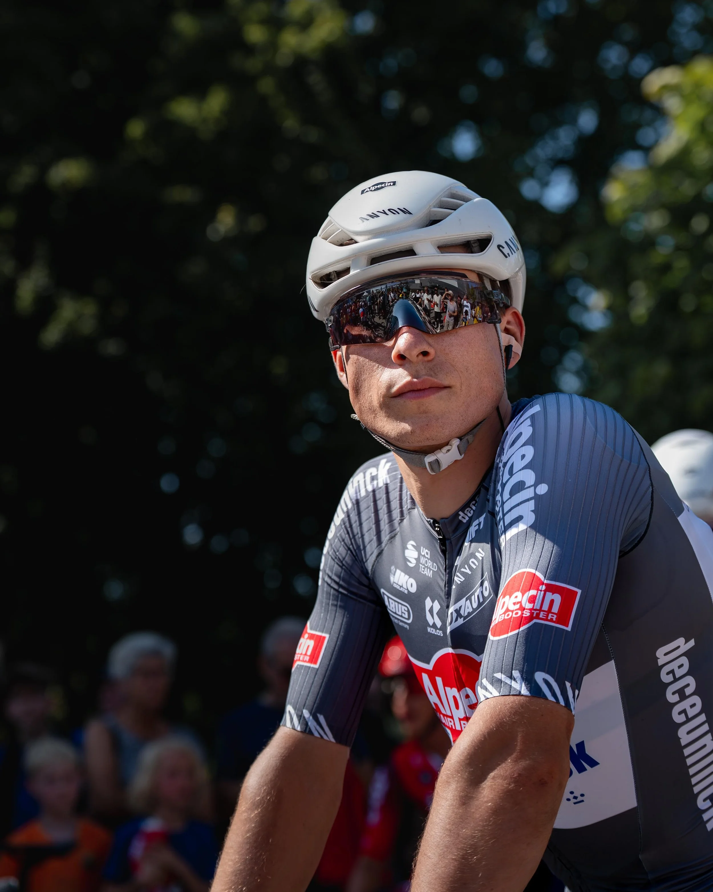 Cykelrytter i sportsudstyr med hjelm og solbriller, delvist synlig, ved start af cykelløb, med publikum i baggrunden.