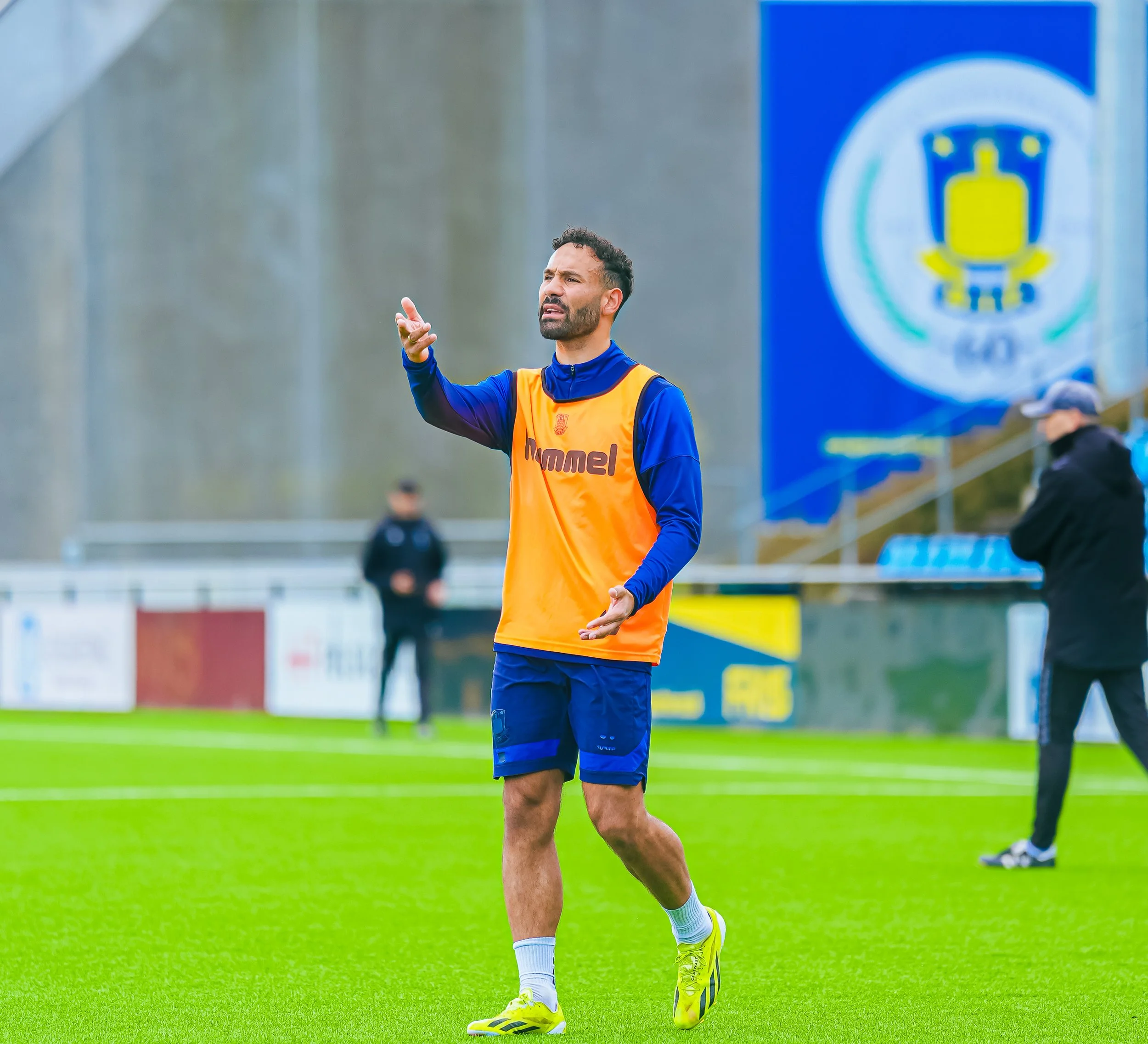 En mand i sportstøj, der står på en grøn fodboldbane, gestikulerer med hånden og har mørkt, krøllet hår og skæg, med en blå trøje og orange vest, og bag ham er en stor blå banner med en logo, og nogle personer i baggrunden.