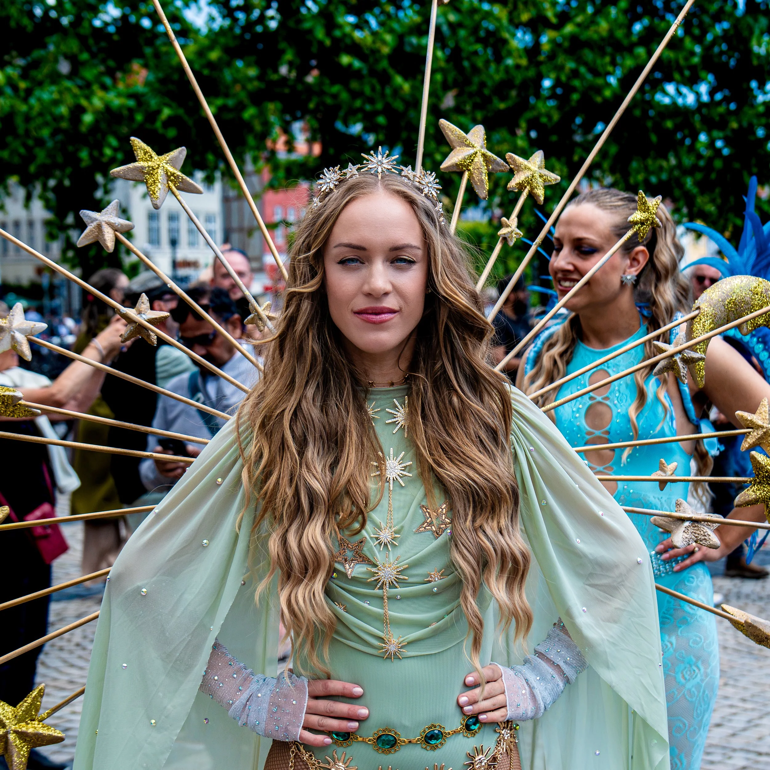 Kvinde klædt i grøn dragt med glitter og stjernehovedbånd, omgivet af folk og pynt i en udendørs parade.